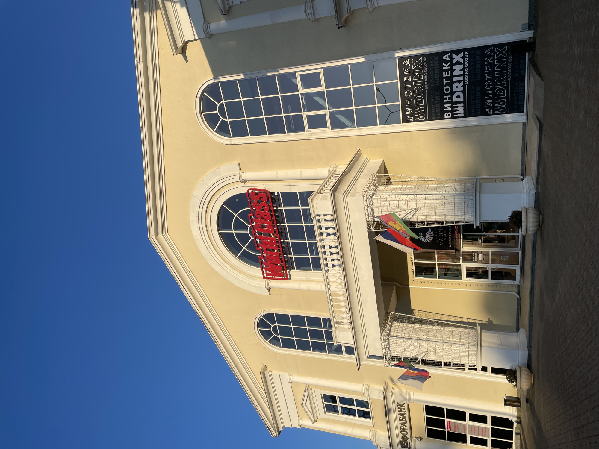 This is a photograph taken at eye level, with the camera tilted slightly upwards. The scene depicts the facade of a light yellow building with classical architectural elements, set against a clear, bright blue sky. The building features large, arched windows with white mullions. Above one of the windows, a red neon sign with white lettering is prominently displayed, but the text is partially obscured.
On the right side of the building, a tall, narrow black banner is visible, with the text "ВИНОТЕКА MDRINX LUDING GROUP" repeated vertically in white lettering. This banner is attached to a glass door or window. Flags are also visible, including what appears to be the Russian flag, mounted on a balcony.
Further down the building, more windows and architectural details are visible, including a balcony with white wrought-iron railings and potted plants. Text visible on a sign at the bottom of the building reads "ФОРАБАНК".
The sunlight casts sharp shadows, indicating that it is likely a sunny day and possibly late afternoon or early morning due to the angle of the light. The overall impression is one of a commercial establishment, possibly a shopping gallery or a bank, in a sunny, urban environment. No people are visible in the image.