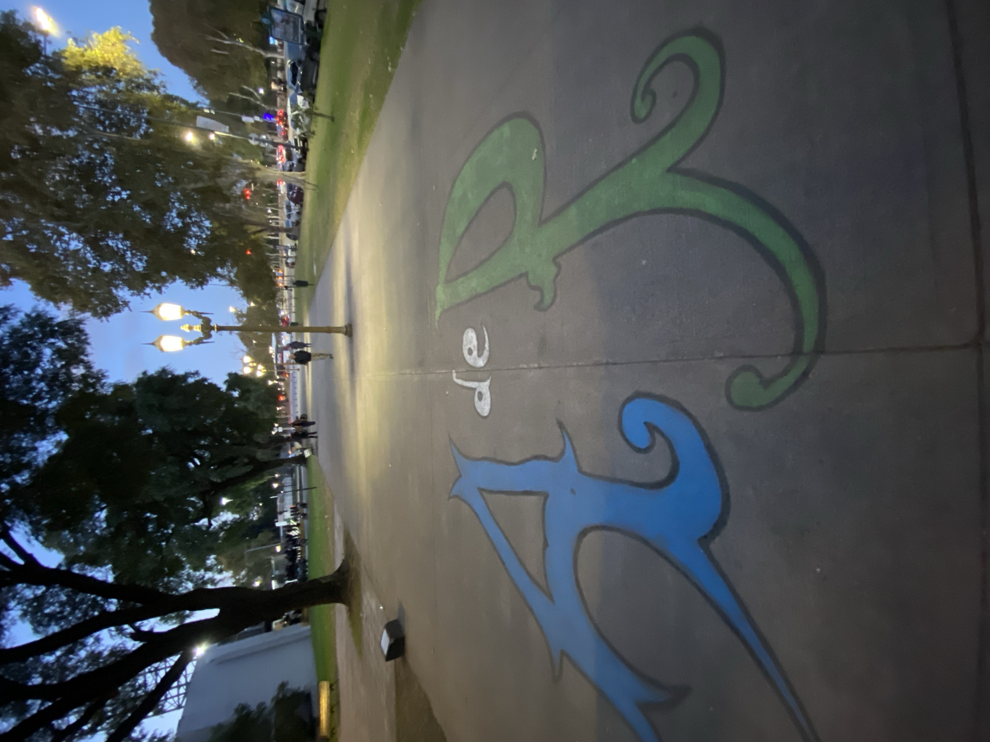 The image shows a sidewalk in Buenos Aires, Argentina, with the letters "BA" painted on the concrete. The "B" is painted in green and the "A" is painted in blue. The image is taken at dusk or dawn, as indicated by the soft lighting and the presence of streetlights. In the background, there are trees lining the sidewalk, and there are people walking along the path.