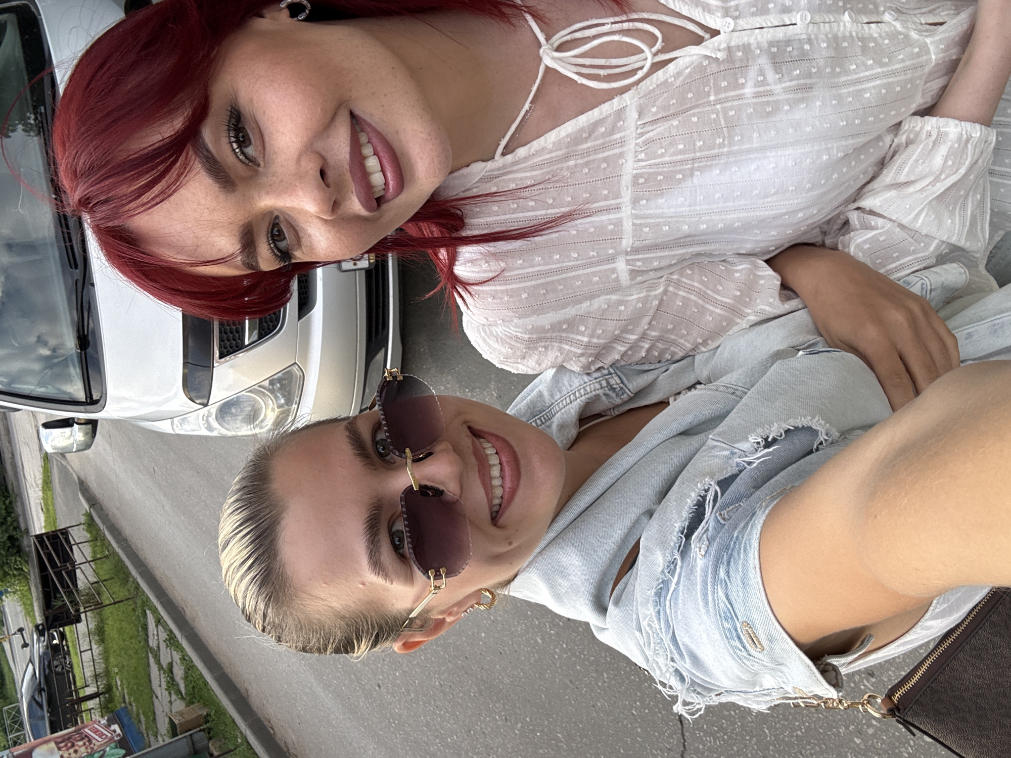 In this selfie, two women are positioned outdoors in what appears to be a city street. The woman on the left has blonde hair pulled back and is wearing sunglasses and a light blue denim jacket. She is holding a dark brown purse. The woman on the right has bright red hair and is wearing a white patterned blouse. They are both smiling and looking towards the camera. In the background, a silver van is visible, parked on the side of a paved road. There is also a sidewalk, grass, and other vehicles in the distance, suggesting an urban or suburban setting.