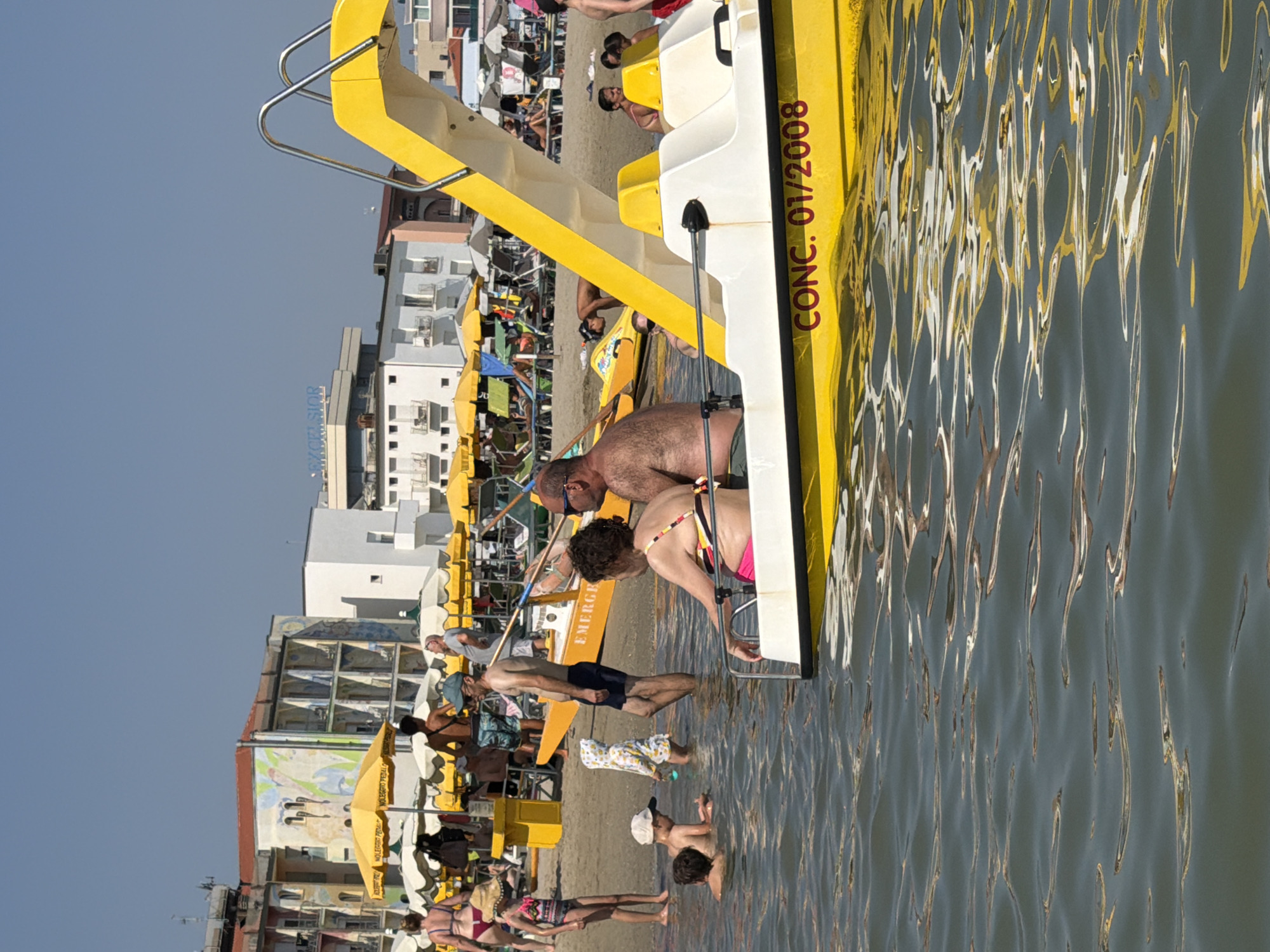 In Gatteo, Italy, on a bright, clear day, a busy beach scene unfolds. The foreground features a bright yellow pedal boat with the designation "CONC. 01/2008" printed on its side. A woman in a pink bikini and a man with a hairy chest are seated within the boat, partially submerged in calm, clear water. Other people are visible in the water, including children, with one child in a hooded outfit. In the middle ground, the sandy beach is populated with numerous yellow and white beach umbrellas providing shade for loungers. Buildings with balconies line the background, some adorned with murals. A few people are walking or sitting on the beach, and others are in the water. The scene suggests a typical summer day at a seaside resort.