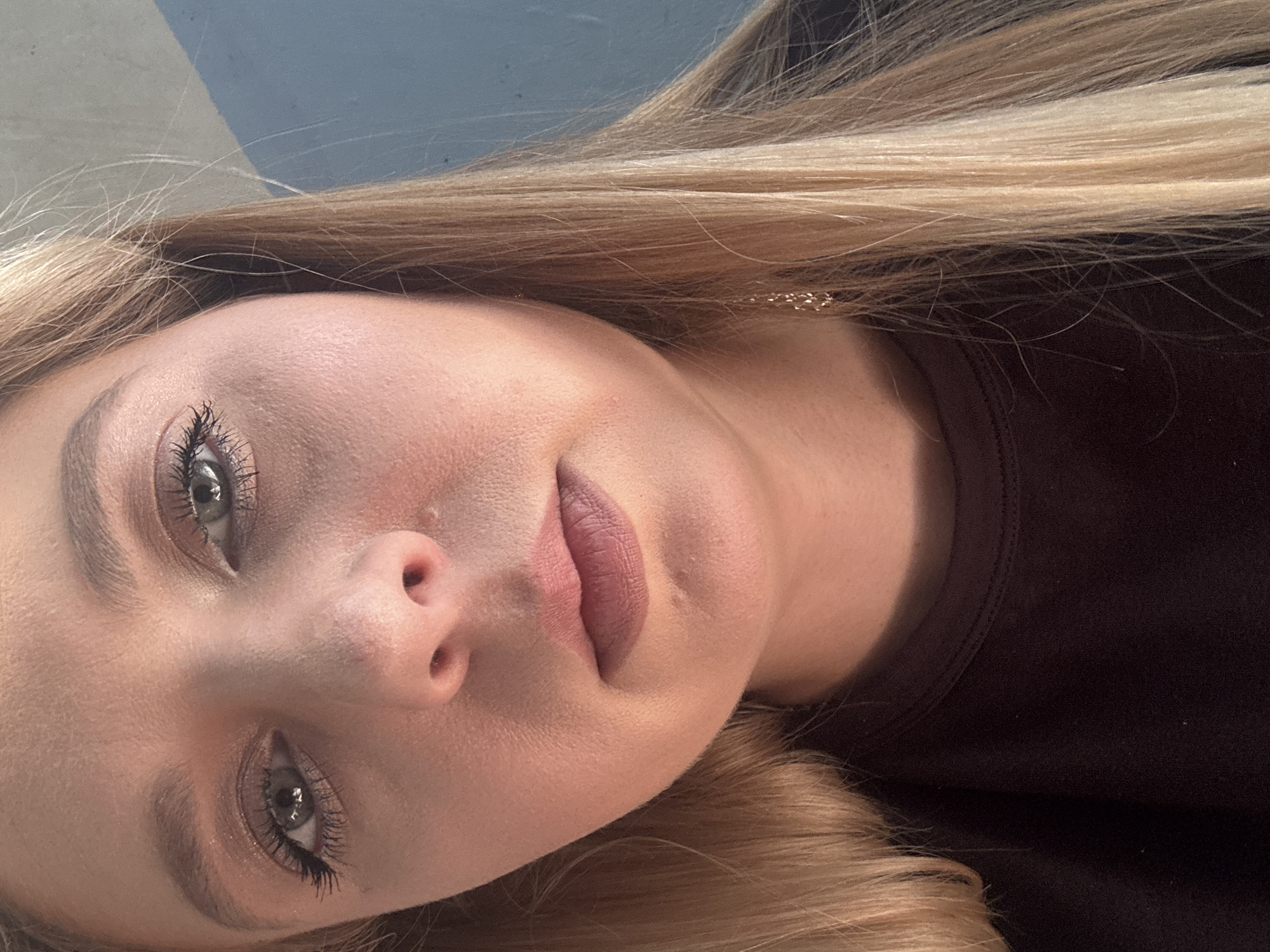 A close-up, overhead selfie of a young Caucasian woman is captured from the chest up. The woman has blonde hair that frames her face and falls over her shoulders. Her eyes are open, and she is wearing makeup, including mascara and eyeshadow. Her lips are parted, and she is wearing lipstick. She is wearing a dark-colored top. A portion of a blue wall is visible in the background, with a diagonal stripe of white light. The lighting suggests the sun is shining from the left, casting subtle shadows on her face. The overall image is a self-portrait taken indoors.