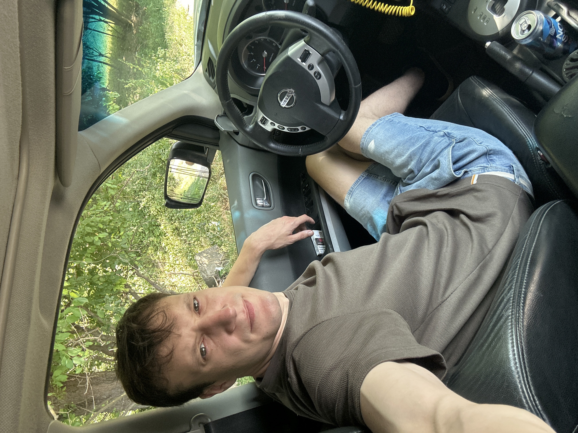A man is sitting in the driver's seat of a car, facing the camera. He is wearing a brown t-shirt and blue denim shorts. His left arm is resting on the door panel, and his right hand is on the steering wheel. The car's interior is visible, including the dashboard, steering wheel, and gear shift. Outside the car, green trees and vegetation are visible. The image appears to have been taken during the daytime. The specific location in the city of Makiv, Ukraine is not discernible from the image.