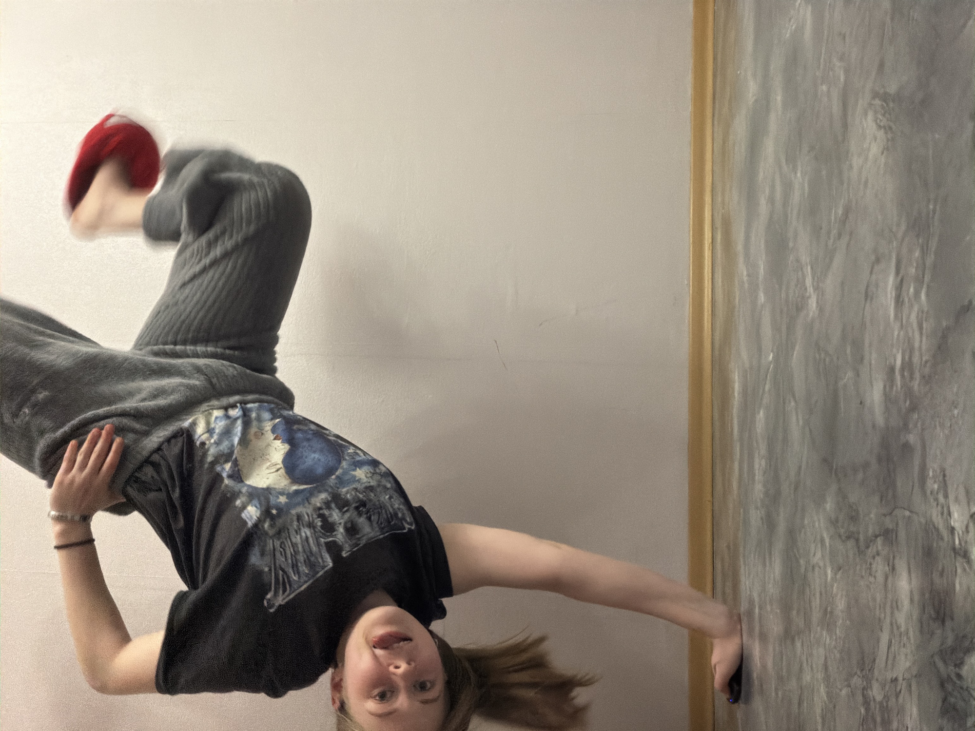 The image presents a young individual captured in an inverted, dynamic pose, as if performing an acrobatic move or playfully hanging upside down. The person, likely female, has light skin and light brown hair that cascades downwards towards the bottom of the frame, emphasizing the inverted perspective.
She is wearing a black short-sleeved t-shirt featuring a prominent blue and white graphic design on the chest, possibly depicting a celestial body like a moon or planet surrounded by intricate patterns. Her lower body is clad in dark grey, ribbed sweatpants. On her left wrist, she wears both a black and a silver-toned bracelet. A blurry red object, possibly a sock or a small item, is visible on one of her feet, with its motion blur suggesting active movement.
Her face, visible in the lower left portion of the image, shows a playful expression with a slightly open mouth and her tongue subtly extended, looking directly at the camera. Her right hand is gripping a textured grey wall, which runs vertically along the right side of the frame and displays streaks of lighter tones, hinting at a concrete or decorative finish. A distinct golden-brown vertical trim separates this textured wall from a plain, light off-white wall that forms the majority of the background. Her left arm is bent, contributing to the balance of her pose.
The setting appears to be indoors, within a room or studio, given the distinct wall types and lack of external elements. There are no discernible cues for the time of day or weather. The overall impression is one of energetic engagement or a moment captured during an active, inverted posture. This media file was captured in Perm, Russia.