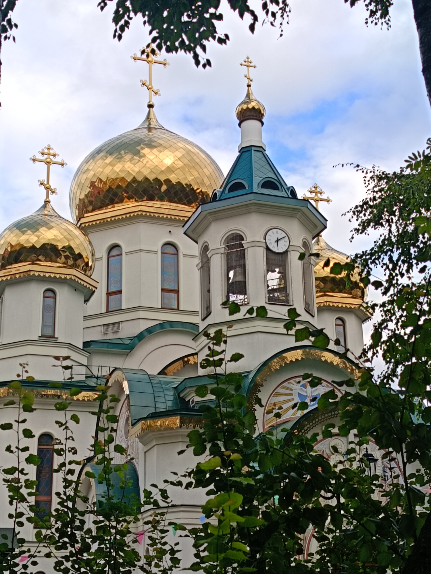 This is a low-angle shot of the Church of the Resurrection of Christ in Saint Petersburg, Russia. The church's white facade is adorned with several golden domes topped with crosses. A prominent clock tower with a teal roof stands to the right of the central dome. Green foliage from trees partially obscures the lower portions of the church. The sky above is cloudy with patches of blue.