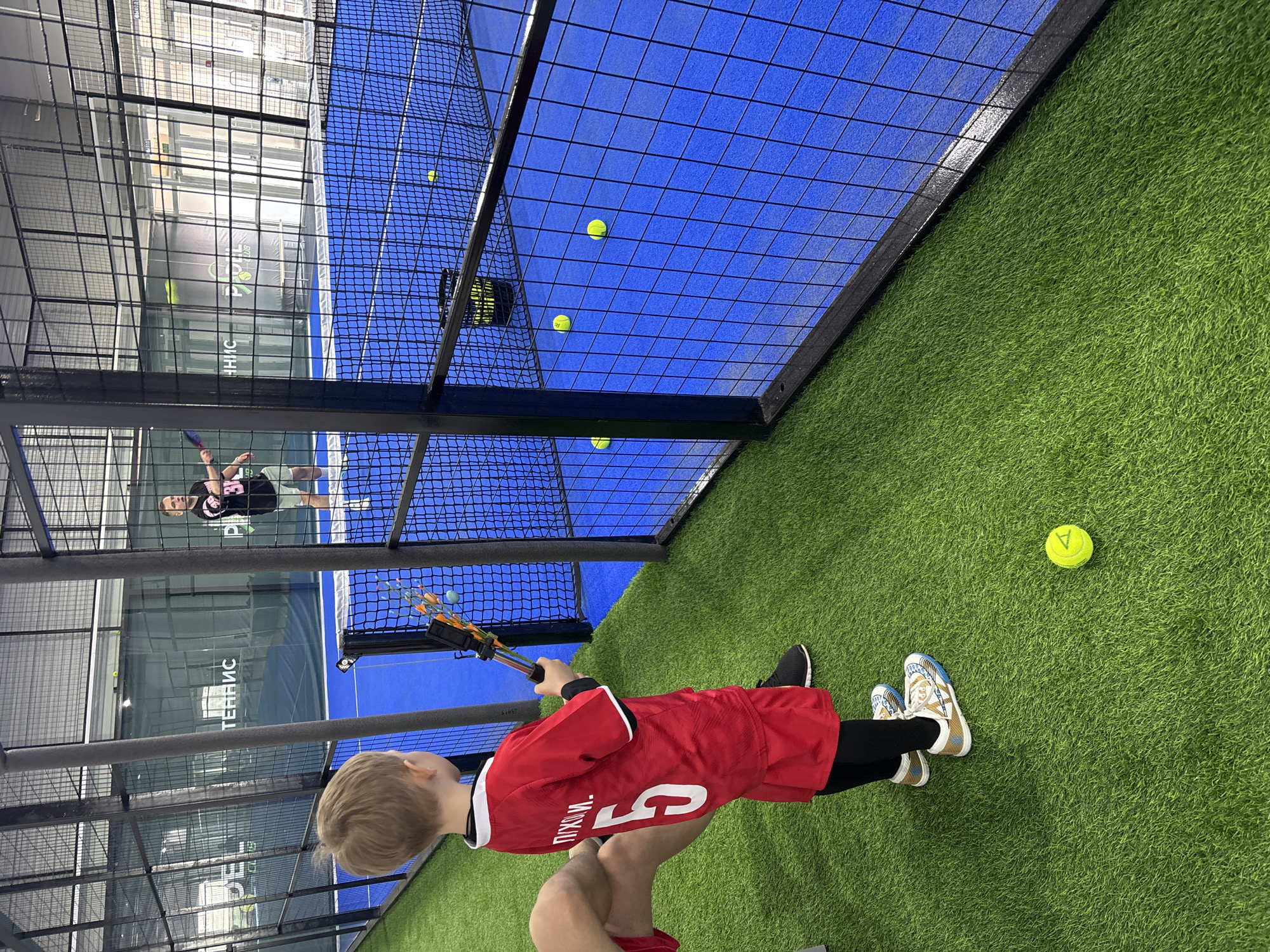 A low-angle shot shows a young boy wearing a red jersey with a white number "5" and the name "ПРИХОМ." in Cyrillic script, holding a ball retriever, standing on artificial green turf. The boy is blond and is looking toward a blue paddle tennis court enclosed by a black wire fence. Several tennis balls are scattered on the court surface and the turf. An adult can be seen in the foreground, holding the child, with partial view of their shorts. In the background, another adult in sports attire is playing on a paddle tennis court with blue walls. The image appears to be taken inside a paddle tennis center in Chaikovsky, Russia.
