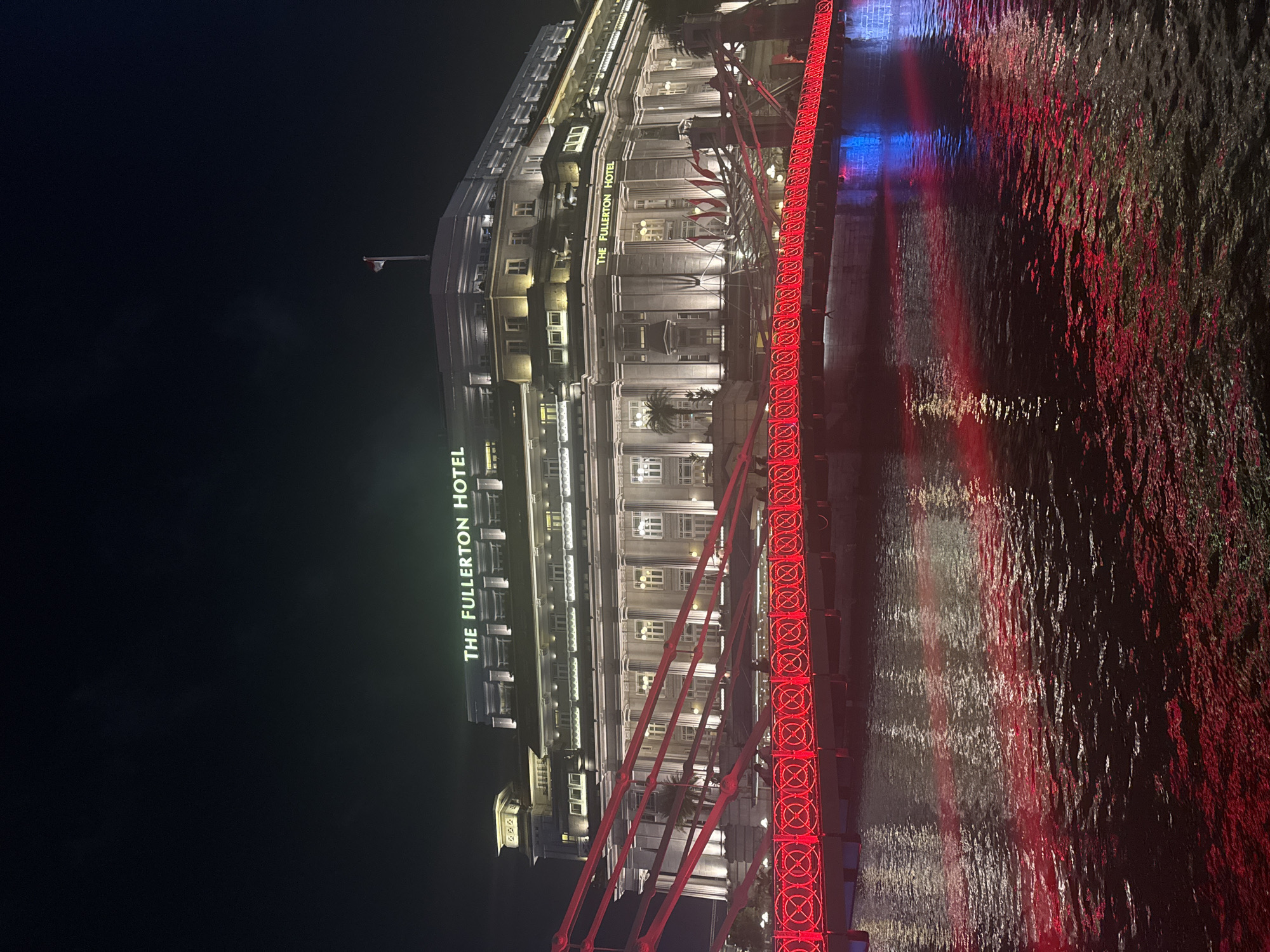 The image depicts a night view of The Fullerton Hotel in Singapore. The iconic hotel, a former post office building, is illuminated, showcasing its grand colonial architecture. In the foreground, a bridge, likely the Cavenagh Bridge or Elgin Bridge, is adorned with bright red lights that form a continuous pattern. These lights are reflected in the dark water of the Singapore River, creating streaks of red and some blue hues. The sky above is dark, typical of nighttime, with the hotel's name "THE FULLERTON HOTEL" prominently displayed in lit-up letters on its facade. A flag, appearing to be the flag of Singapore, is visible atop the hotel. No people are clearly discernible in the image. The overall scene is a vibrant display of urban nighttime scenery in Singapore.