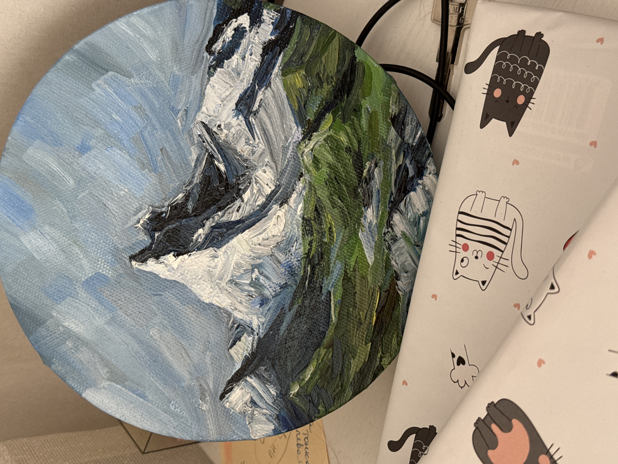 The image shows a close-up of a circular painting of a mountain landscape. The left side of the painting depicts a sky with shades of blue and white. The right side features a mountain with green, white, and gray tones, suggesting vegetation and snow. Beside the painting is a wrapped gift with a cat theme. The cats are cartoon-like and in various poses, with pink hearts scattered on the white wrapping paper. A metal frame of some kind is also visible. It is not possible to determine from the image if the painting is located in Krasnoyarsk, Russia or any other location.
