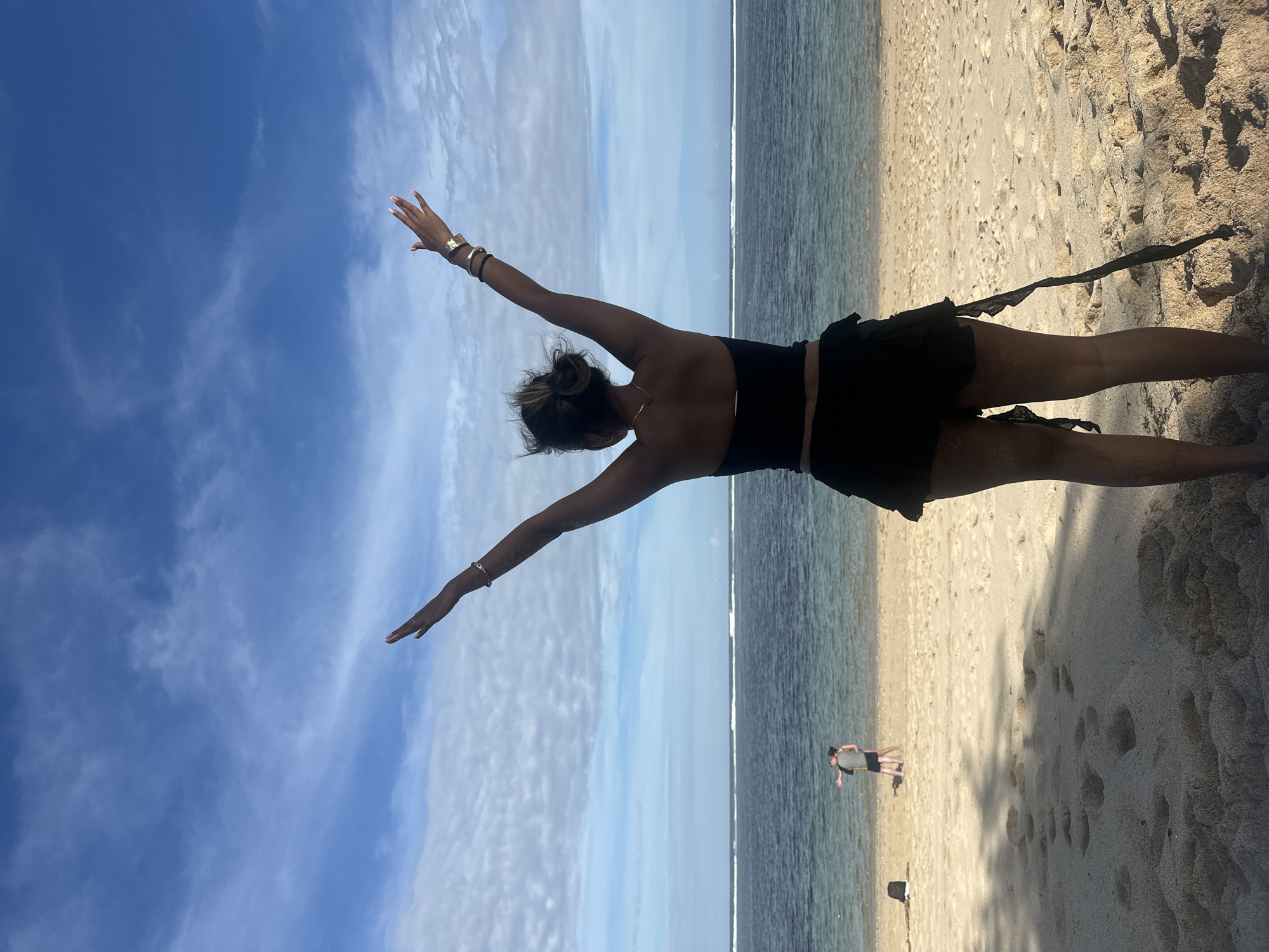 This image depicts a person performing a dynamic pose on a sunny beach in Kuta Selatan, Indonesia. The main subject, seen from behind, is a person with their arms outstretched in a balanced posture, resembling a yoga pose, looking towards the ocean and sky. They are wearing a black top and black shorts or a skirt, with gold-colored bangles on their wrists and their hair tied back in a bun.
The setting features a bright blue sky with wispy white clouds above the vast expanse of the ocean. The foreground and right side of the image show light-colored sand with subtle textures, leading to the calm, turquoise waters of the sea on the left. A faint line of waves is visible along the distant shoreline. In the mid-ground, a smaller figure of another person can be seen standing near the water. The bright, clear lighting indicates it is daytime, likely a sunny morning or afternoon. There is no visible text in the image.