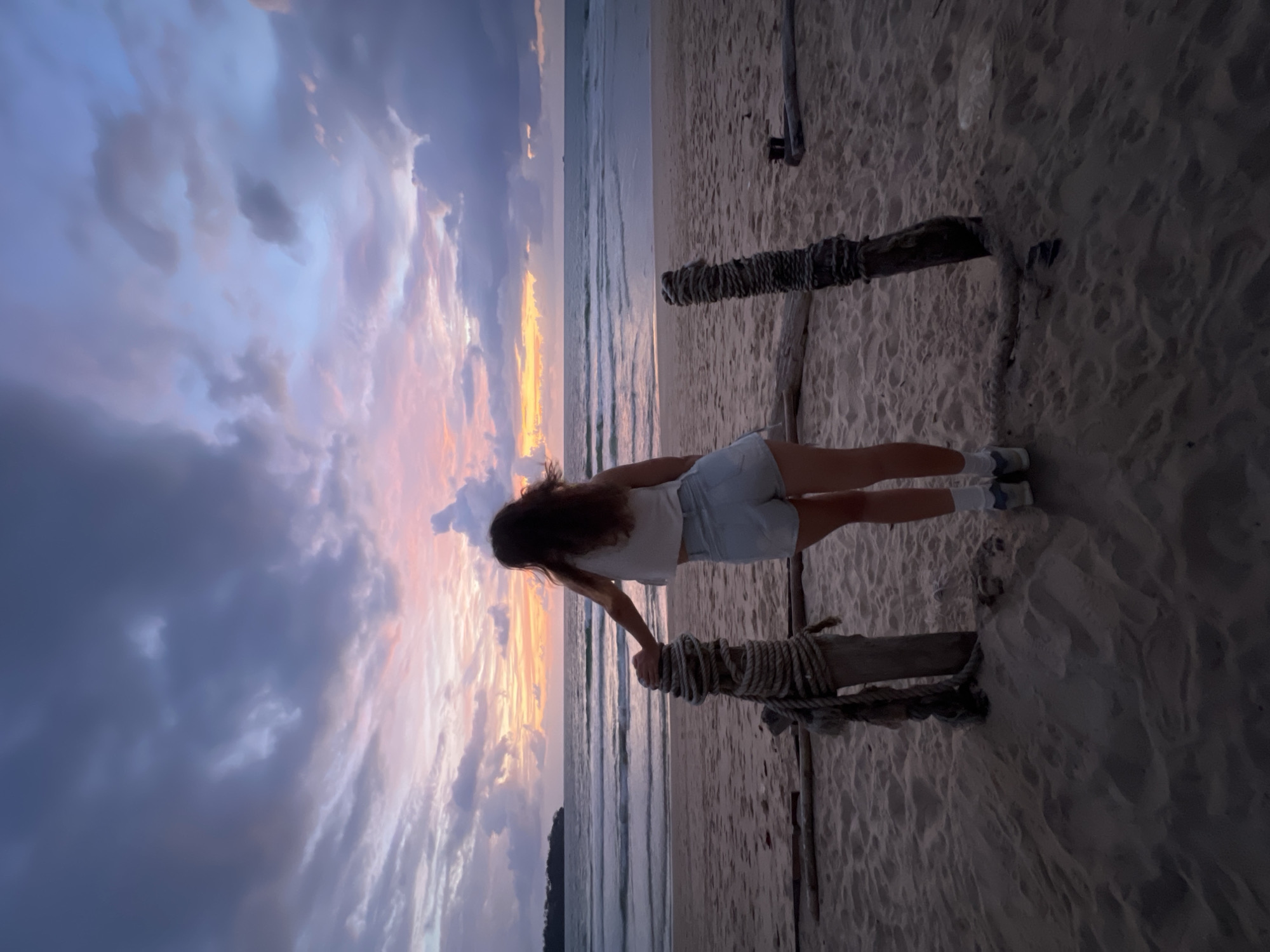 A woman stands on a beach in Choeng Thale, Thailand, facing the ocean at sunset. She is viewed from behind, wearing a white tank top and denim shorts, with her left arm resting on a wooden post wrapped in rope. The post is part of a weathered wooden barrier on the sand. The ocean is calm with gentle waves, and the sky is filled with dramatic clouds colored by the setting sun in shades of pink, orange, and purple. The sand is a muted brown, showing footprints and indentations. The overall scene depicts a serene, atmospheric moment at dusk.