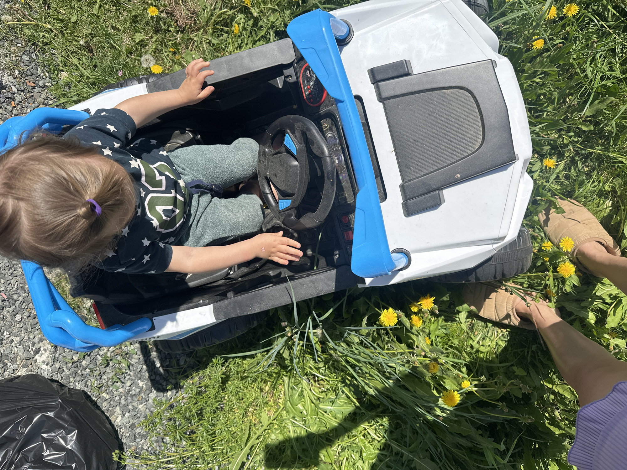 This overhead shot captures a young child seated in a white and blue toy car, positioned outdoors in Kharnavy, Russia. The scene is bathed in bright sunlight, indicating daytime.
The child, viewed from above, has light brown hair tied with a purple elastic band. They are wearing a dark blue t-shirt adorned with white stars and a prominent green and white design on the back, along with grey trousers. Their hands rest on the black steering wheel, which has blue accents, suggesting they are actively playing or preparing to drive the toy vehicle.
The toy car features a predominantly white body with distinct blue roll-bar-like structures or handles on either side. The dashboard is black with various dials and buttons, including red and silver elements. A black panel on the side of the car exhibits a simulated carbon fiber texture.
The setting combines patches of green grass densely populated with bright yellow dandelions and areas of grey gravel. To the lower left, a black trash bag is visible on the gravel. In the upper right and along the right edge of the frame, an adult's feet, clad in light brown slippers, are seen standing in the grass among the dandelions, alongside a portion of a purple garment. This indicates an adult is nearby, likely supervising the child. No clear, legible text is visible in the image.