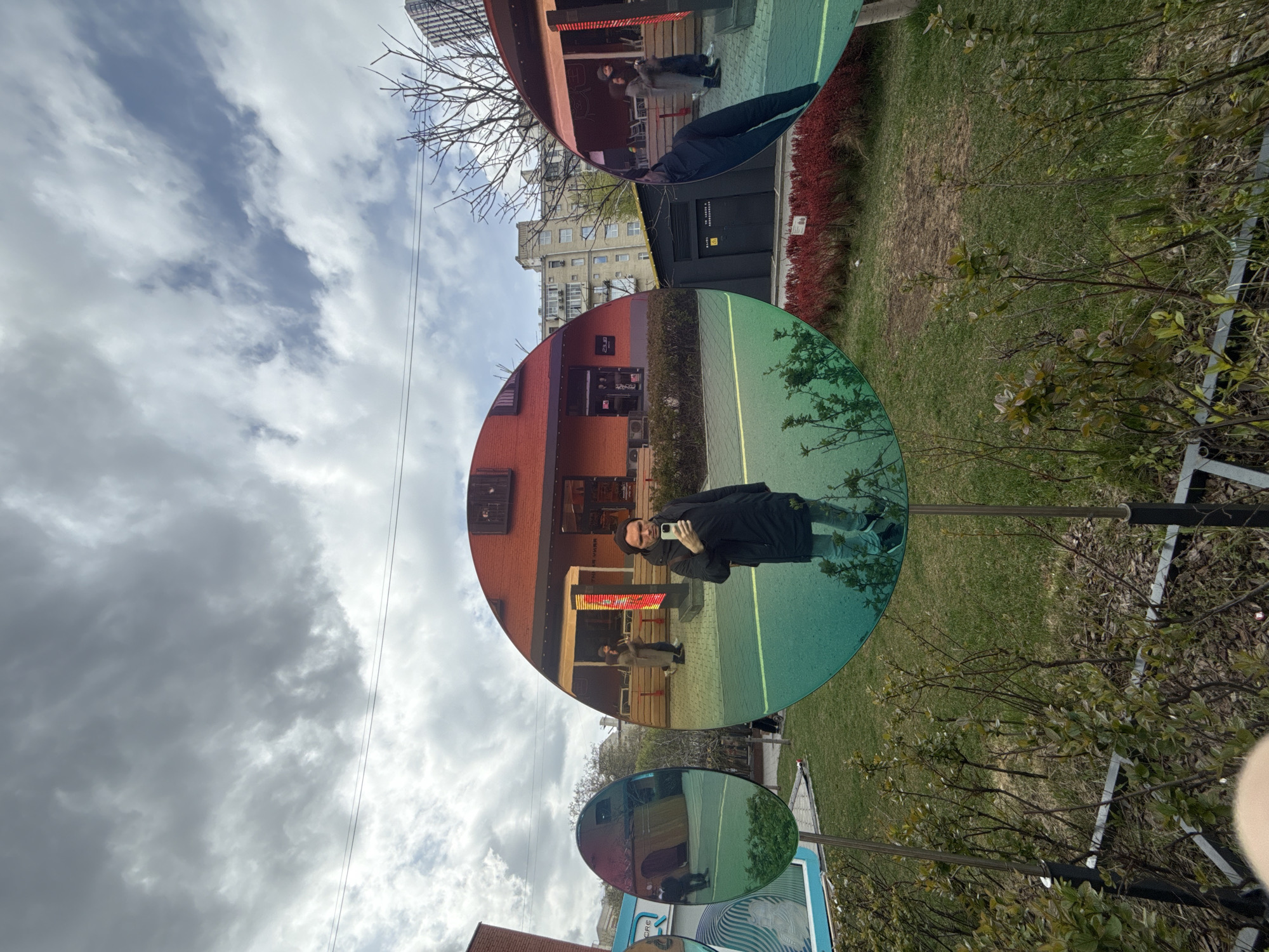 The photograph shows a person taking a picture of themselves in a spherical public art installation in Moscow, Russia. The artwork consists of three iridescent, circular mirrors on metal stands placed on a grassy lawn adjacent to a sidewalk. The mirrors reflect parts of the surroundings, including red brick buildings, trees, and a cloudy sky. A couple of people are reflected in the top mirror.