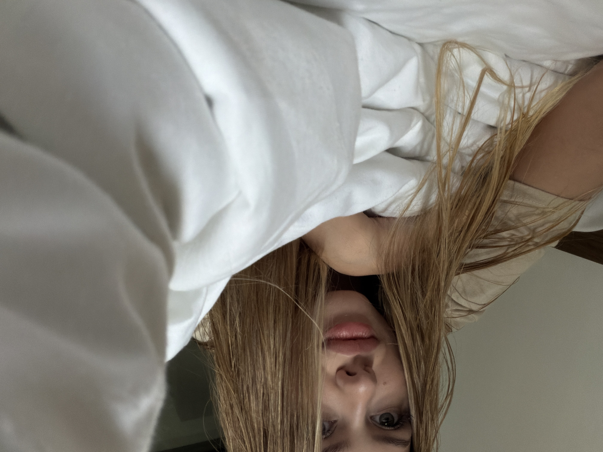 An extreme close-up, low-angle, selfie-style shot shows a young Caucasian woman with long, dirty blonde hair lying on a white bed, partially covered by white sheets. Her head is tilted slightly, and she is looking at the camera with a neutral expression. Her lips are full and appear to be slightly parted. Her skin is pale, and she is wearing a light-colored t-shirt. The background consists of a plain, light-colored wall and a dark, out-of-focus area that could be furniture or part of a room. The overall lighting is soft and diffused, creating a natural, intimate atmosphere. The image provides no specific clues or indicators of its location within the city of Đà Nẵng, Vietnam, or any other identifiable urban environment.