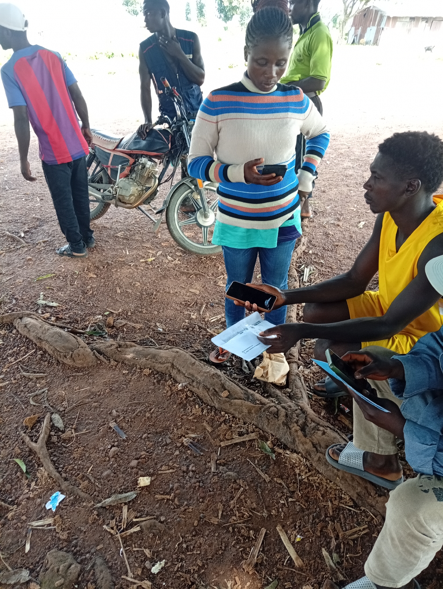 The image depicts a group of people gathered outdoors in Fadan Kuower, Nigeria. In the foreground, a woman wearing a striped sweater and jeans is holding a smartphone. To her right, a man in a yellow sleeveless shirt is squatting and looking at a document. Another person in a blue denim jacket is partially visible, also looking at a tablet. Behind them, a man in a blue vest and a young man in a colorful t-shirt stand near a motorcycle. The ground is covered in dirt, small rocks, and exposed tree roots. In the background, vegetation and a simple structure with a corrugated metal roof are visible. The overall scene suggests an informal gathering possibly related to business or community activity.