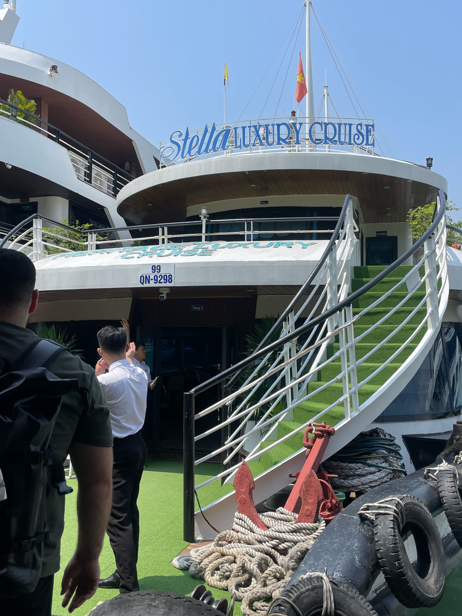 The image shows the exterior of a luxury cruise ship named "Stella Luxury Cruise" docked in Quang Ninh City, Vietnam. The ship is white with blue lettering on the upper deck. A red and yellow flag flies from a mast. A green carpeted staircase with white railings leads up to the ship's entrance. Two men are visible in the foreground, one in a dark shirt and backpack, and another in a white shirt and black pants. Another man in a uniform is standing by the ship's entrance. The ship is docked on a body of water, indicated by the presence of ropes and an anchor in the foreground. The sky is clear and blue.