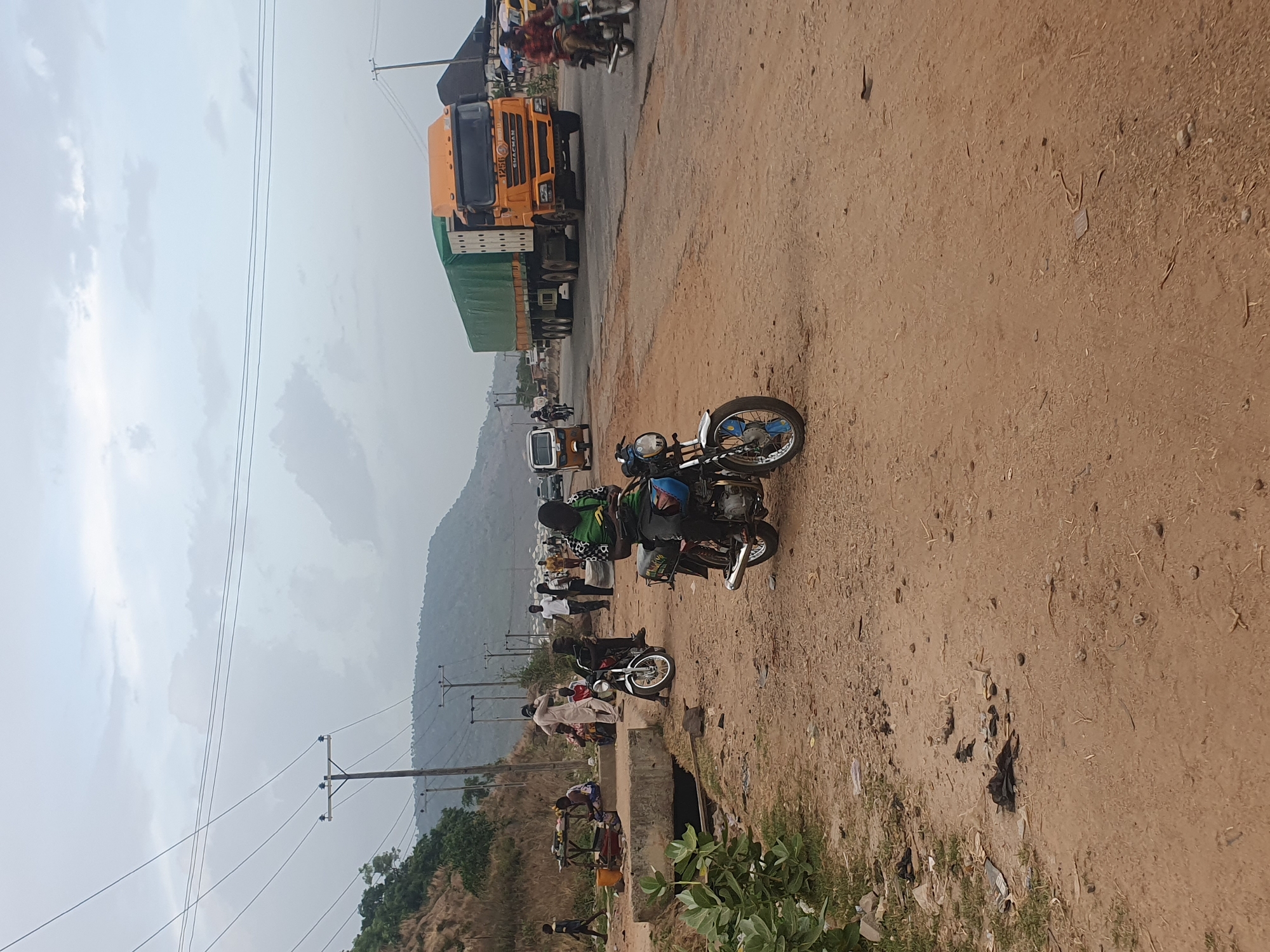 This media file captures a vibrant roadside scene in what appears to be a semi-urban or rural area, consistent with the provided location context of Lokoja, Nigeria. The image is taken from a slightly elevated perspective, looking across a dusty, unpaved shoulder towards a paved road, with hills and an overcast sky in the background.
In the immediate foreground, the dusty, reddish-brown ground is visible, showing scattered debris and sparse vegetation. A motorcycle is parked or idling on the dirt shoulder, with a rider wearing a green and black patterned shirt and a blue helmet.
The midground features several people and vehicles. More motorcycles are visible, some with riders or passengers. One three-wheeled auto-rickshaw is also present further down the road. People are standing and walking along the roadside, suggesting a daily routine of transport and commerce. A concrete culvert or drain runs parallel to the road, separating the dirt shoulder from a slightly lower area with more vegetation. Power lines strung between wooden utility poles traverse the scene.
On the paved road in the background, a large yellow and green articulated truck dominates the view. The truck bears the brand name "SHACMAN" and the number "1256" on its front grille. Other vehicles, including more motorcycles, are visible around the truck. Beyond the road, large, green-covered hills rise under an overcast or hazy sky.
The activity suggests a busy transport hub or stop along a main route. The weather appears to be cloudy daytime. No specific emotions are discernible among the people, who seem engaged in their daily activities. The scene is characterized by a mix of infrastructure, human activity, and natural landscape.