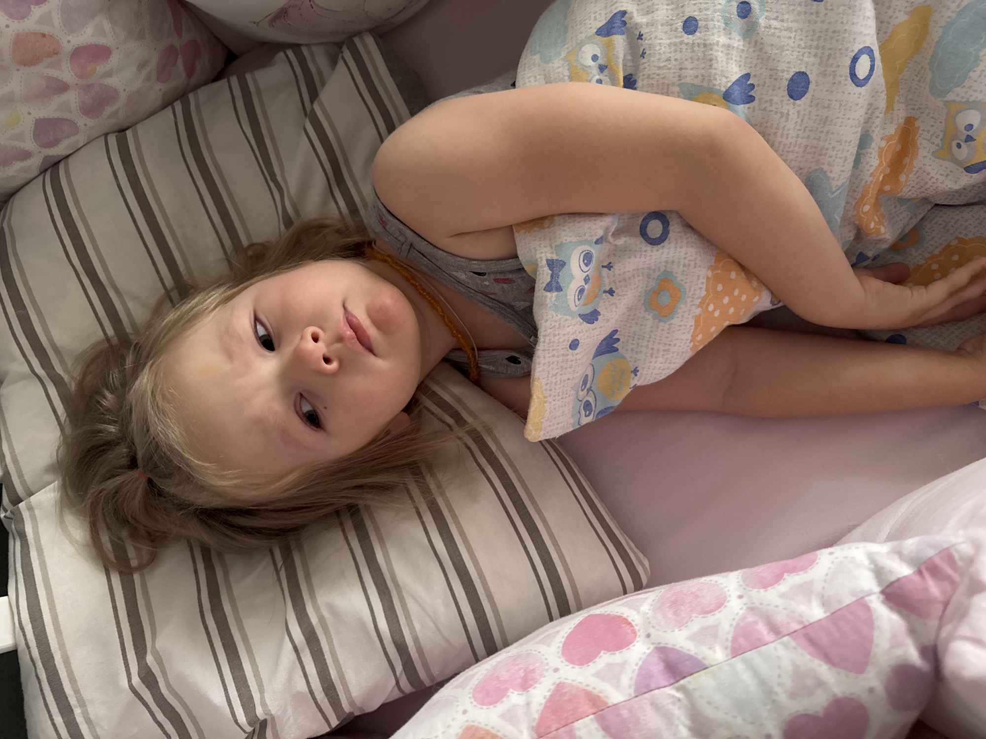 A young child with blond hair lies in a bed. The child is under a blanket patterned with owls and geometric shapes. The bed has several pillows, including one with a stripe pattern and another with hearts. The child wears a gray tank top and a necklace with amber-colored beads. The location is implied to be Gornyi (Dvurechenskoe S/P), Russia. No other details are discernible about the setting.