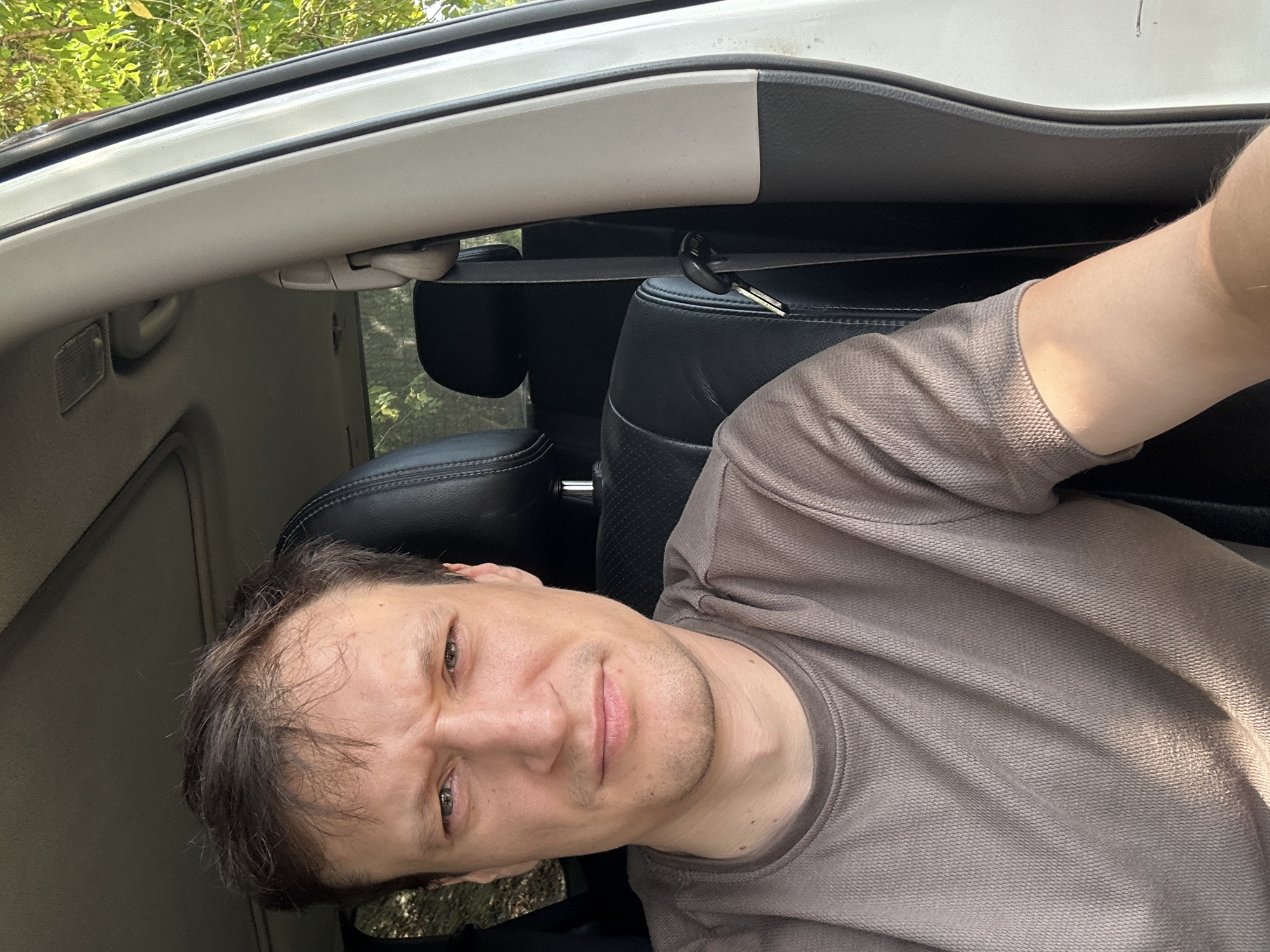 A close-up, forward-facing selfie is captured from within a vehicle. The subject, a light-skinned male with brown hair and a furrowed brow, is positioned in the driver's seat. He is wearing a taupe-colored t-shirt. The interior of the vehicle, with its black leather seats and white trim, is visible. Sunlight filters in from the right, illuminating the subject's arm and chest. The exterior, characterized by green foliage, suggests an outdoor setting, potentially in the city of Makiv, Ukraine, given the prompt's context. The image is taken during daylight.