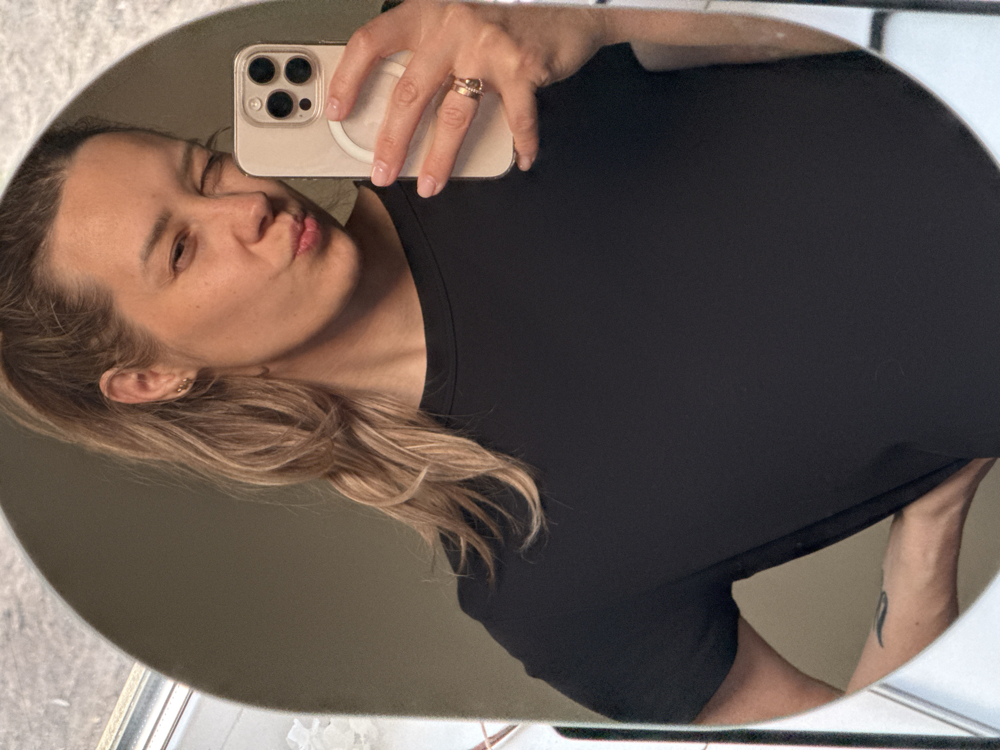 A woman is taking a selfie in a mirror. She is wearing a black t-shirt and has blonde hair pulled back in a ponytail. She is pouting her lips for the camera and holding a smartphone with three cameras. The phone case is beige. She is wearing rings on her fingers. The mirror is oval-shaped and has a reflective surface. Her left arm is slightly visible, revealing a black tattoo near her elbow. The background is blurred. Based on the mirror and surrounding textures, it is likely she is in a bathroom, possibly in a personal residence. There is nothing in the image to indicate the location is Chaikovsky, Russia.