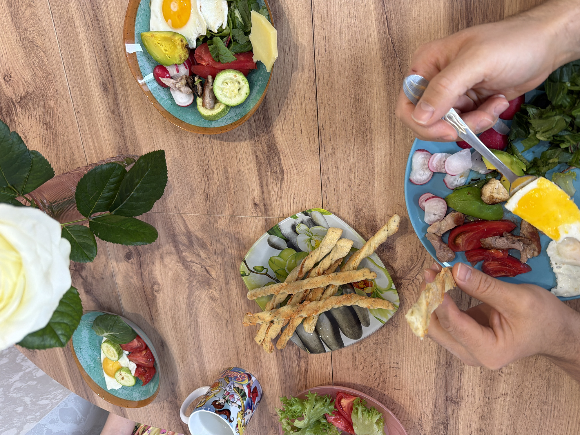 The image shows a top-down view of a wooden table spread with food. There are four plates with various salads, including greens, tomatoes, radishes, zucchini, avocado, cheese, and fried eggs. One plate also contains meat. A plate of breadsticks is also visible. A person's hands are present, holding a piece of breadstick and a spoon. A vase with a white rose, and a mug with a cartoon design are also on the table. The location in the city of Izhevsk, Russia is not discernible from the image.