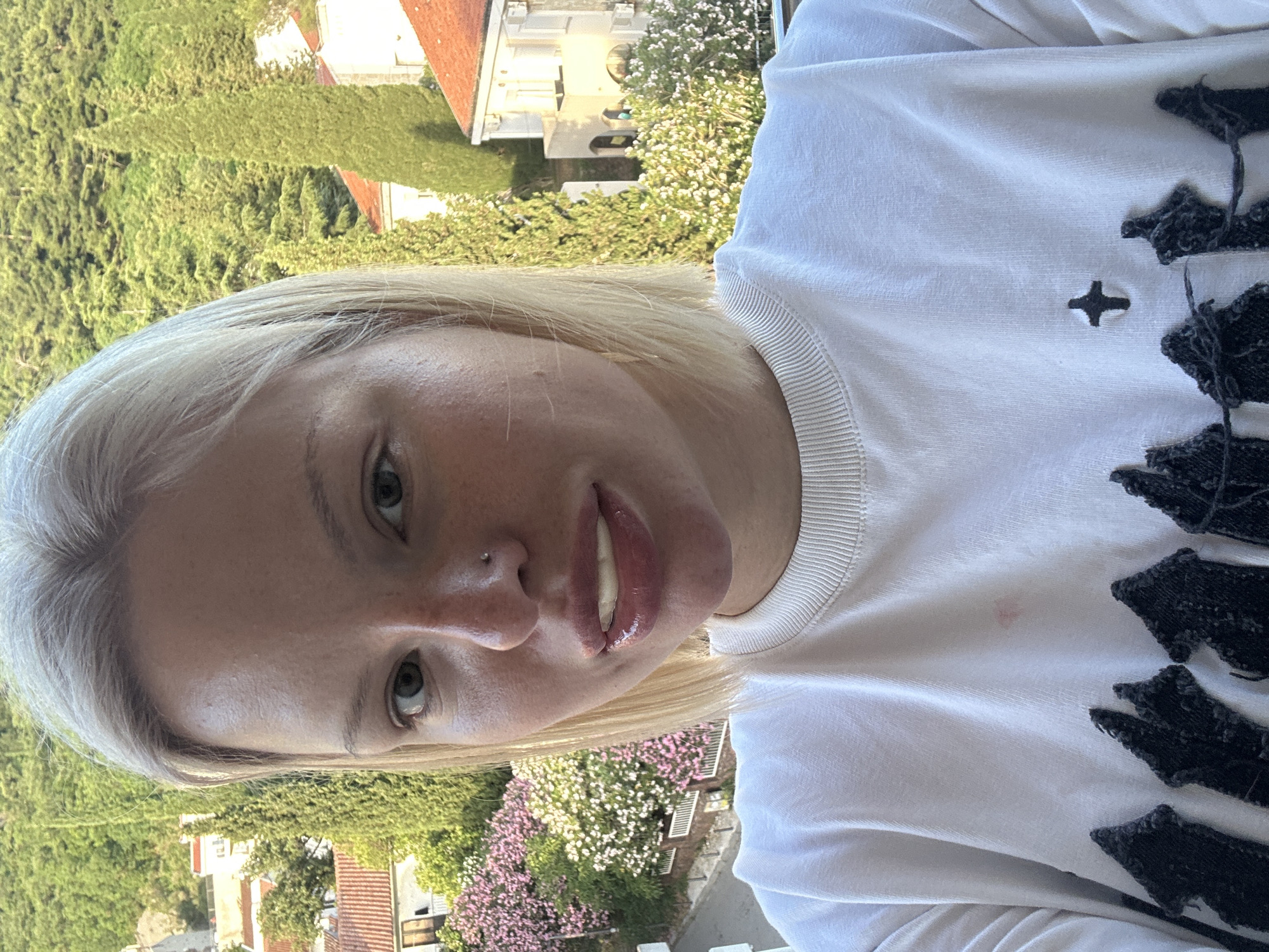 The image shows a fair-skinned woman with blonde hair, capturing a selfie from an upward perspective. She looks directly at the camera with a neutral to slightly smiling expression. Her eyes are light-colored, and she has a small silver nose piercing. Her lips are glossy and slightly parted, revealing some teeth. She is wearing a white long-sleeved top or sweatshirt with a ribbed collar. The garment features distinct black, abstract embroidered elements on the viewer's right side, along with a small black, four-pointed star-like symbol on the left chest area. A faint reddish mark is noticeable on her upper left chest.
The background, visible above the woman's head and shoulders, portrays a sunlit outdoor landscape. It is dominated by dense green foliage, including trees and what appears to be a manicured hedge. In the distance, several buildings with classic terracotta roofs and white facades are visible, interspersed with clusters of white and pink flowering plants. The bright daylight indicates a clear, sunny day. This picturesque setting is consistent with the specified location context of Budva, Montenegro. No visible text is present.