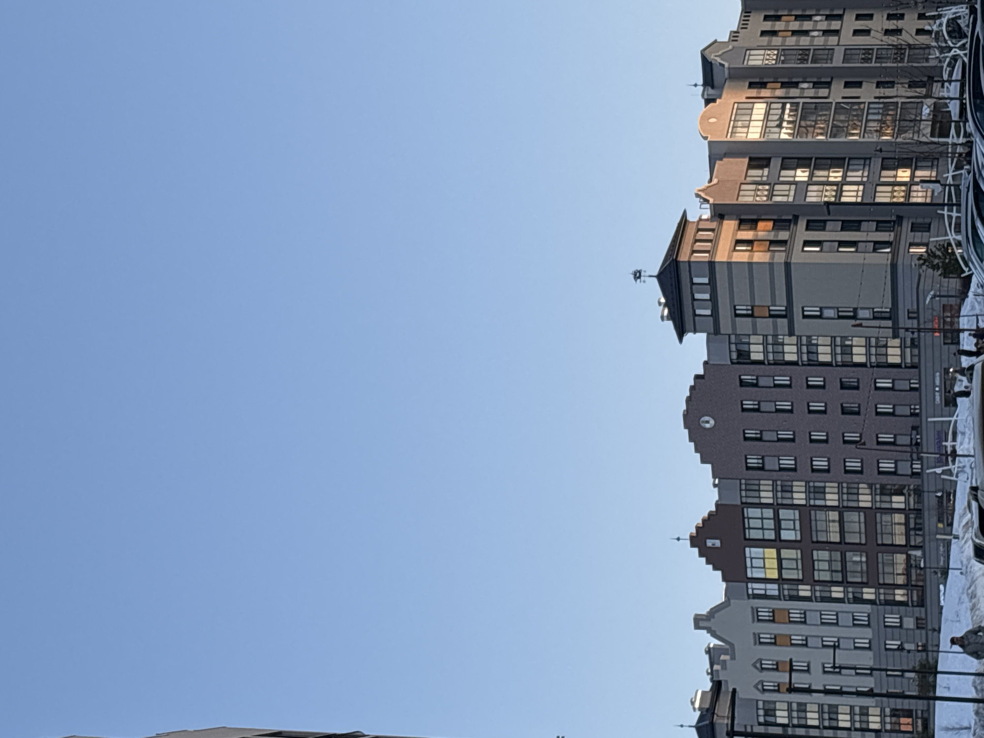The scene depicts a modern urban environment in Izhevsk, Russia, specifically the "Stary Amsterdam" residential complex at Kirova Street, 109. The upper two-thirds of the image are dominated by a clear, light blue sky under bright daylight conditions. The lower third features a row of seven multi-story buildings, approximately 6-8 floors tall, characterized by varied facades in shades of grey, reddish-brown, and light brown, and distinct gabled or stepped rooflines, evocative of Northern European architecture. Numerous windows, many reflecting direct sunlight with an orange hue, are visible across the building fronts. One building, the third from the left, features a prominent square tower-like structure topped with a dark peaked roof and a rooster weathervane. The fifth building from the left exhibits a clock face near the peak of its stepped gable.
The ground in the foreground is covered in a substantial layer of white snow. Several dark-colored cars are partially visible, parked in what appears to be a parking area, partially obscured by snowdrifts. Streetlights with multiple luminaires are spaced along the base of the buildings. In the lower-left quadrant, several indistinct human figures are present on the snow-covered ground; one individual in a light-colored jacket is moderately visible, walking towards the right. Commercial signage, including "КАФЕ" (Cafe), is visible on the lower levels of some buildings. No distinct interactions between individuals are discernible.