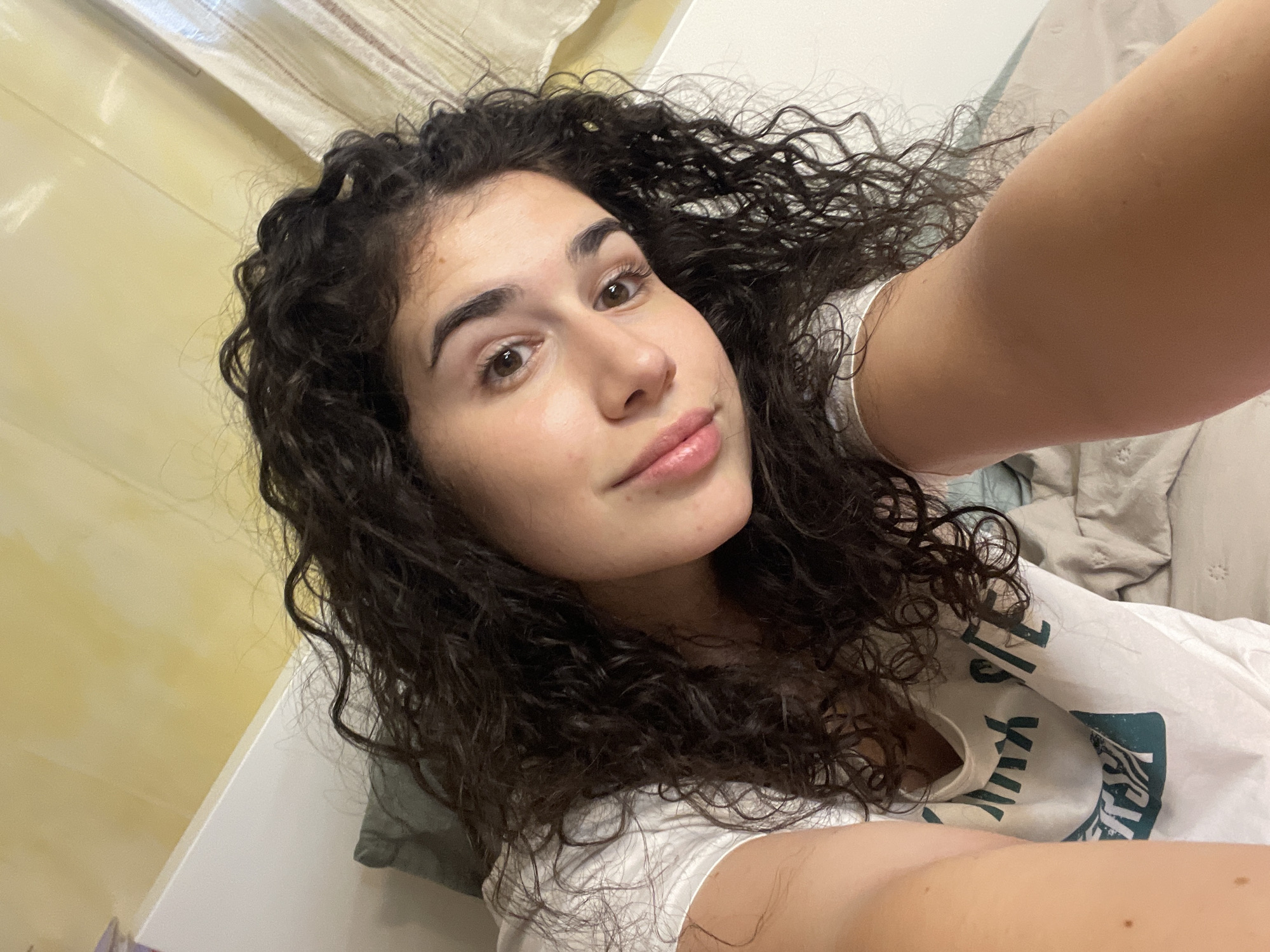A young woman with long, dark, curly hair is pictured taking a selfie, looking directly into the camera with a calm, slightly smiling expression. Her light brown eyes are open, and her lips are slightly parted. She has fair skin.
She is wearing a white t-shirt with a dark green graphic design on it. Partially visible text on the shirt includes letters like "LE" and a stylized "X", along with other indistinct characters or imagery.
The setting appears to be indoors, likely a bedroom. In the background, there's a light-colored wall or headboard, and a light-striped curtain or fabric is visible in the upper left corner. Crumpled light-colored bedding or blankets are also present, suggesting she is reclining or lying down. Her right arm, possibly holding the camera, is visible in the bottom right corner of the frame. The lighting suggests it is daytime or a well-lit interior. The overall atmosphere is relaxed. The location context provided is Sofia, Bulgaria.