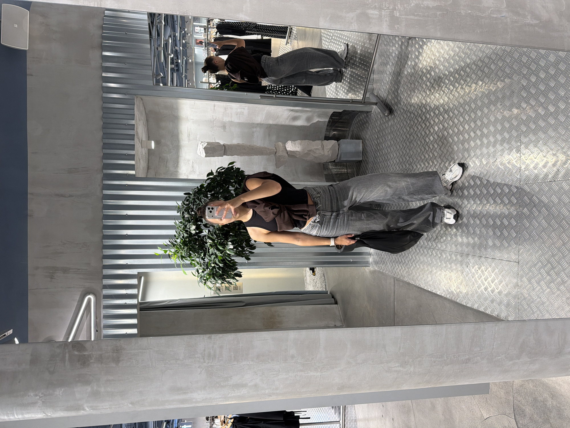 The image shows a young woman taking a mirror selfie in a retail store. She is wearing a dark sleeveless top, grey wide-leg pants, and white sneakers. She is holding a black bag in her left hand and her phone in her right hand, using it to take the photo. A green potted plant is positioned in front of her, partially obscuring her legs.
The setting appears to be a modern, minimalist boutique. The walls are a mix of concrete and corrugated metal. A large mirror dominates the scene, reflecting the woman, the plant, and the interior of the store. In the reflection, racks of clothing, some with polka dot patterns, are visible, suggesting a clothing store. The floor is made of diamond plate metal.
The lighting is bright and even, indicating it is daytime, likely indoors. There are no discernible weather cues. The overall mood is casual and observational, with the woman appearing relaxed.
While no explicit text is visible on the woman's clothing or the immediate surroundings, there might be text on the clothing tags or store signage reflected in the mirror, but it is not legible in this image. The location is specified as Moscow, Russia.