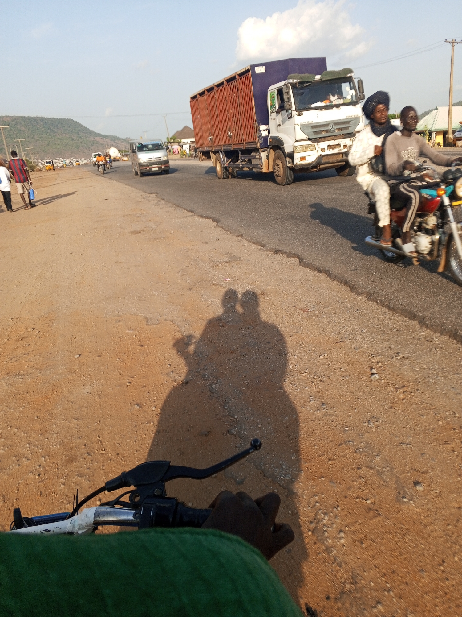The image, likely taken in Lokoja, Nigeria, shows a street scene from the perspective of someone riding a bicycle. The foreground displays the bicycle's handlebars and the rider's hands, with a green cloth partially visible. The shadow of two people on a motorcycle is projected onto the ground. On the right edge of the road, two people ride a motorcycle past a large truck. Several people walk alongside the road and other vehicles drive by, including a light-colored van. In the distance is a low mountain. The sky is bright with some clouds.