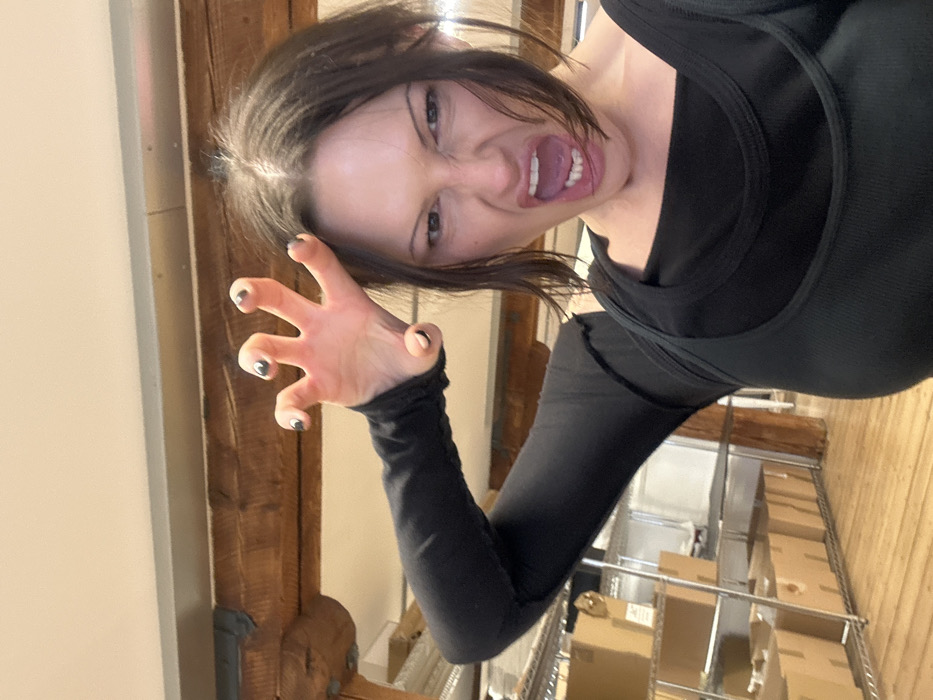 An indoor, medium-shot image captures a single female individual performing a mock-aggressive pose directly facing the camera. The subject has dark hair and wears a black long-sleeved top underneath a dark, likely black or dark green, ribbed tank top. Her left arm is raised with the hand open, fingers splayed and curled, mimicking a claw gesture. Her fingernails are painted dark with lighter tips. Her face displays a "growling" expression, characterized by a wrinkled nose, narrowed eyes, and an open mouth revealing upper teeth. The background features exposed architectural elements including a prominent rough-hewn wooden beam running horizontally, with a metal bracket visible on the left. Behind the subject and to her left, a metal shelving unit holds numerous cardboard boxes. A wooden floor is visible on the lower left. The lighting is even, suggesting artificial illumination. The specific location within the city of London, United Kingdom, cannot be ascertained from the visual evidence provided.