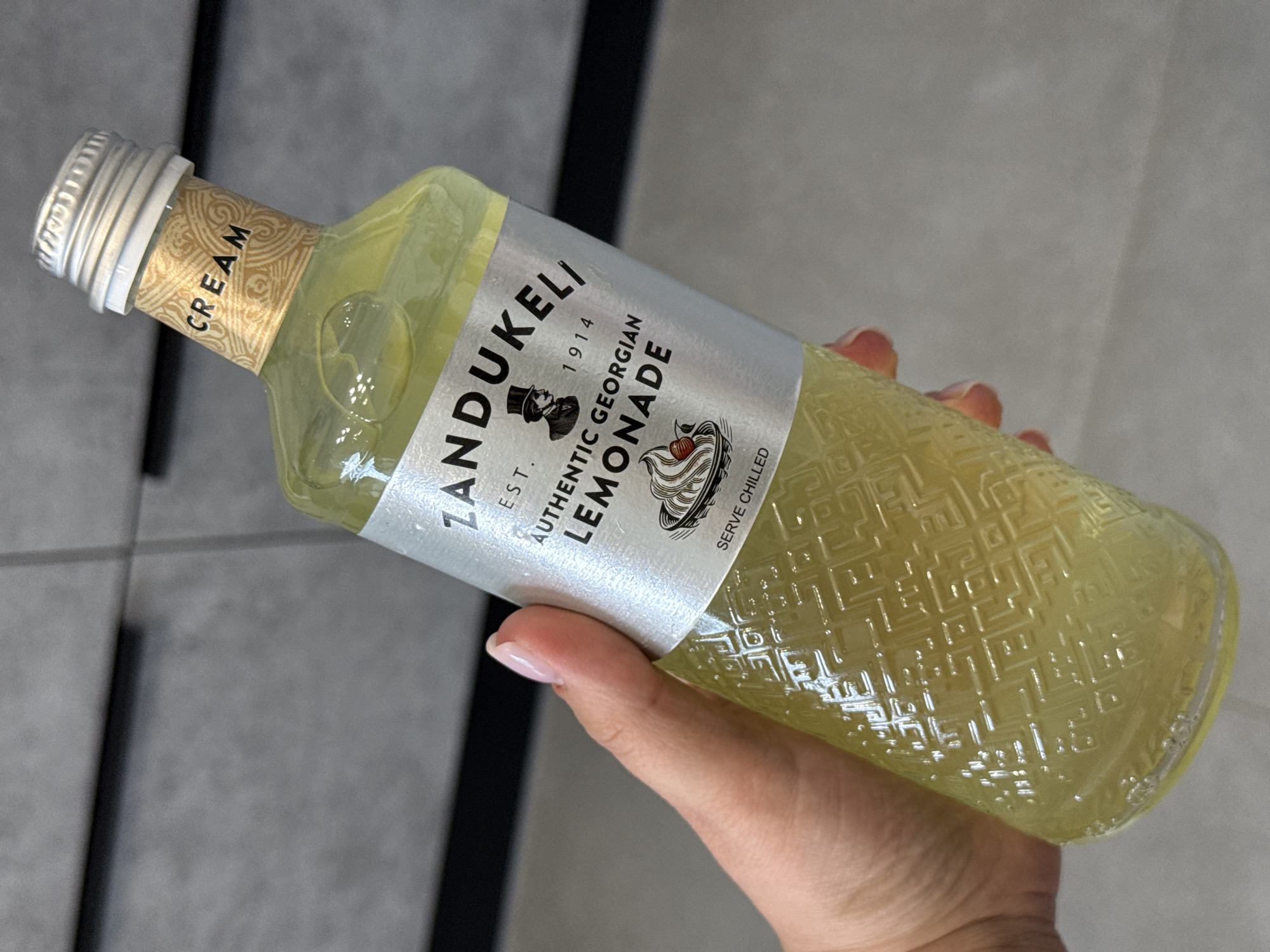 A hand with a light pink nail polish is holding a glass bottle of "ZANDUKELI AUTHENTIC GEORGIAN LEMONADE." The bottle is filled with a pale yellow liquid and has a silver cap. The label on the bottle has a silver background with black text and an illustration of a man in a top hat and a depiction of a dessert. The word "CREAM" is printed in gold lettering on a decorative band around the neck of the bottle. The bottle has a textured pattern on the glass. The background appears to be a grey, textured wall. The image is taken from a slightly high angle.