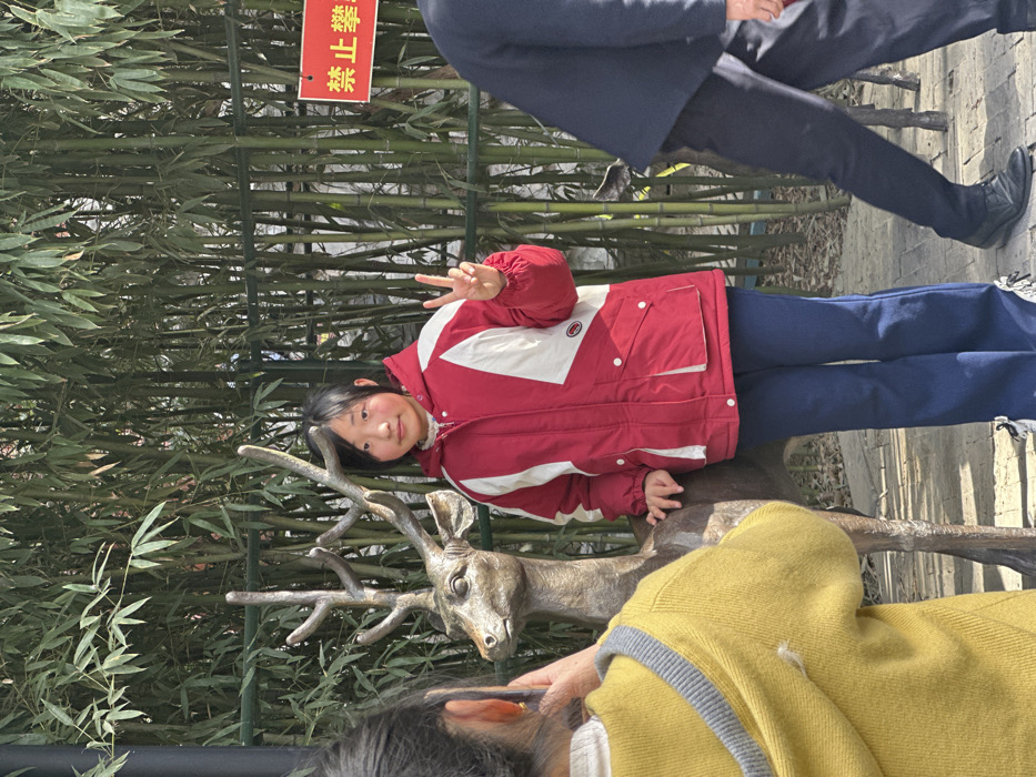 A girl in a red and white coat poses next to a bronze deer statue, flashing a peace sign. Another person in a yellow sweater is partially visible at the bottom of the image. Bamboo stalks form a backdrop, with a red sign bearing Chinese characters affixed to the stalks. An adult figure in a dark suit walks behind them, on a stone walkway.