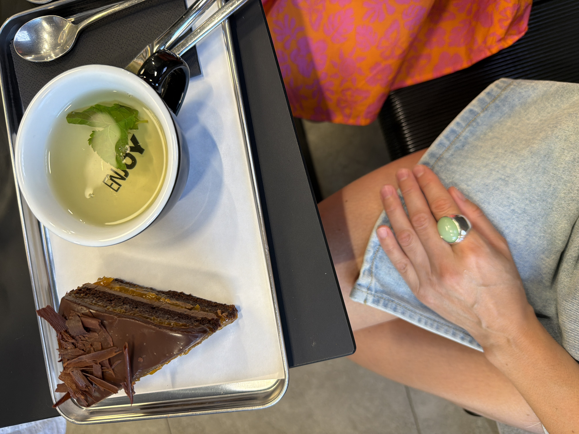 An overhead, medium shot captures a tray holding a slice of chocolate cake with orange filling and a cup of tea with the word "ENJOY" printed on the inside of the cup. The tea contains a mint leaf and appears to be a light yellow color. To the right of the tea is a silver spoon. The tray is resting on a black surface. In the foreground, a person's hands and legs are visible. The person is wearing a light blue denim skirt and a bright orange and pink floral top. The person's right hand is resting on their lap, and they are wearing a silver ring with a large, light green oval stone. The scene is likely indoors, possibly at a cafe or restaurant. There is no information in the image to suggest a specific location within Izhevsk, Russia.