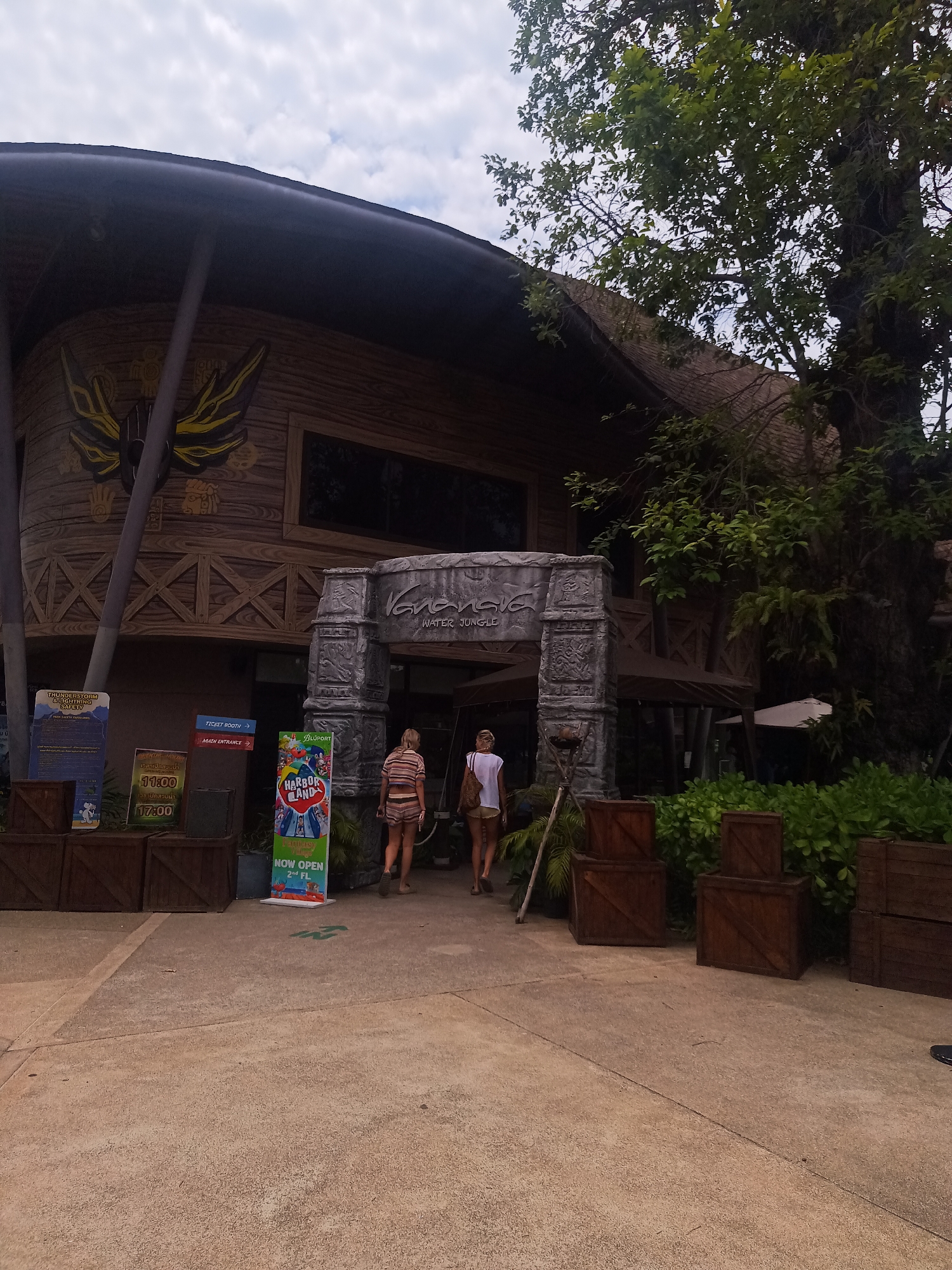 The image shows the entrance to "Vananava Water Jungle", a water park located in Hua Hin City Municipality, Thailand. Two women are walking through the ornate, stone-textured entrance. To the left, signs provide information about thunderstorms and lightning safety, park opening times (11:00 and 17:00), and directions to the ticket booth and main entrance. There's also a sign promoting "Harbor Land" on the 2nd floor of a nearby facility. Wooden crates line the walkway on either side of the entrance. The building behind the entrance has a wooden, jungle-themed facade with a large, stylized eagle-like symbol. A large tree is visible on the right side of the image. The sky is cloudy.