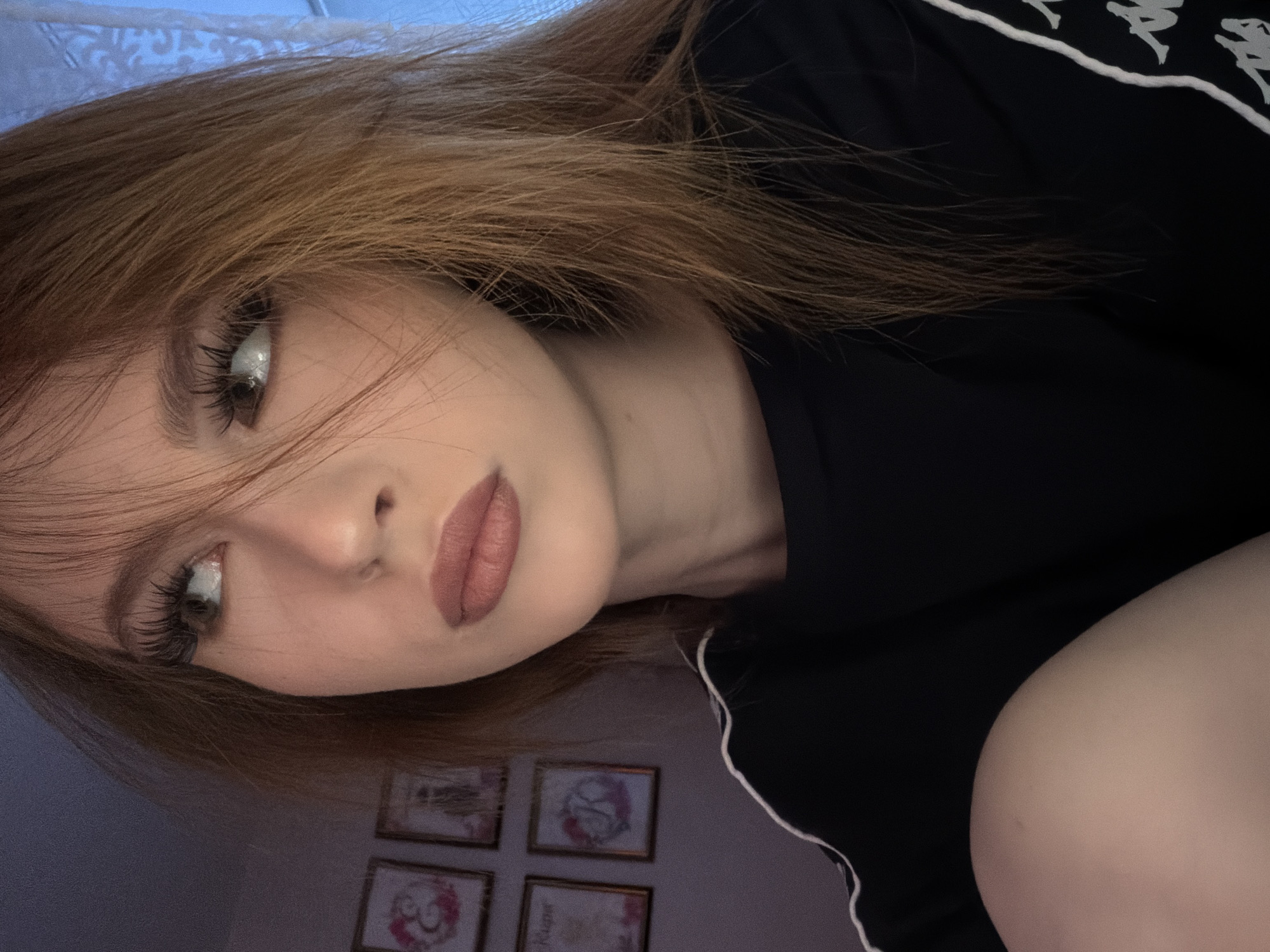 A person with long reddish-brown hair, wearing a black shirt with white stripes, is looking at the camera. The person has fair skin, defined eyelashes, and mauve-colored lipstick. Behind the person, there are four framed pictures hanging on a wall, and partially visible white fabric possibly a curtain. The lighting appears to be from an overhead source. There is no indication of a location in Noviy, Russia in the image.
