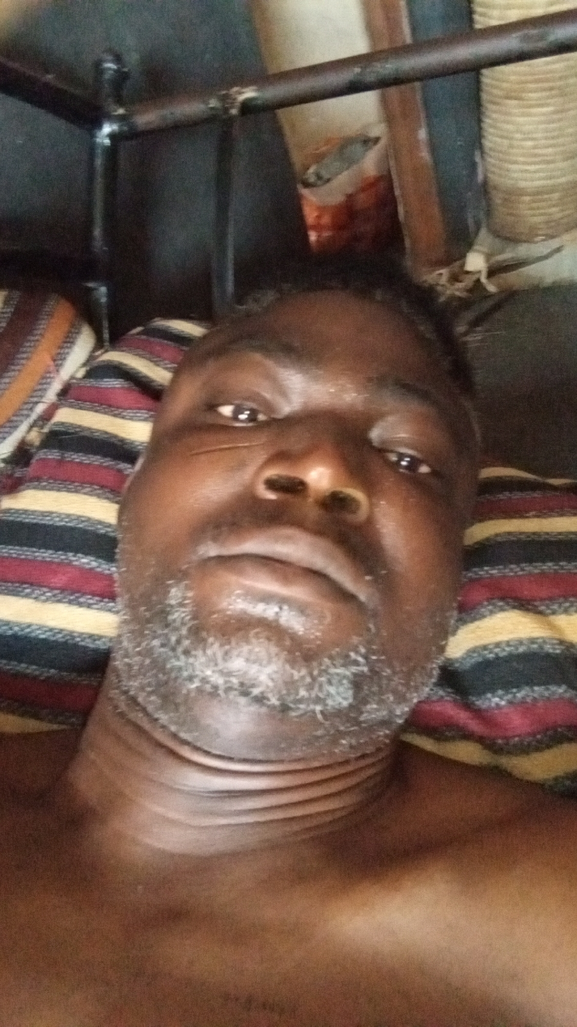 A close-up, low-angle selfie shows a dark-skinned man lying down, looking directly at the camera. He has a short, greyish beard and stubble, and distinct facial scars or tribal marks on his right cheek. His expression is neutral. He appears to be shirtless, revealing folds on his neck. The man is resting on a pillow or blanket with a striped pattern in shades of red, black, yellow, and beige. Above him, a dark metal frame structure is visible, likely part of a bed or bunk bed. In the background, behind the metal frame, indistinct objects can be seen, including what appears to be a stacked woven material or basket, and possibly a container. The setting is indoors with somewhat dim lighting. No text is visible.