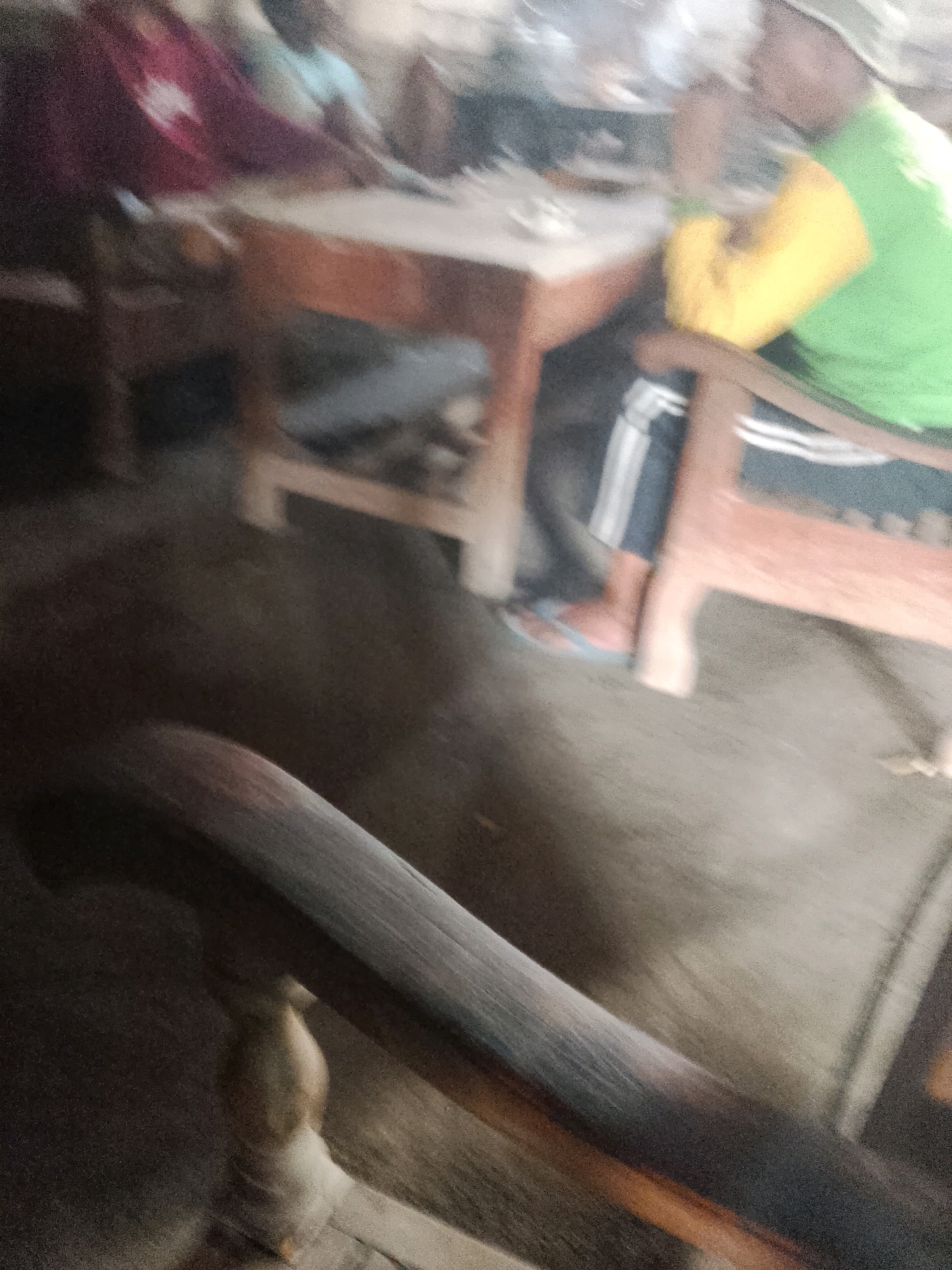 The image depicts a blurred indoor scene, possibly a dining or common area, located in Wadang, Indonesia. Several individuals are present, seated around a wooden table. One person on the right wears a green shirt with a yellow sleeve and black and white striped pants, along with flip-flops. Another individual to the left is partially visible, wearing a maroon garment. The environment appears rustic with a dirt floor and wooden furniture, suggesting a humble dwelling or community space. The overall image is out of focus, making it difficult to discern specific details about the people or their interactions.
