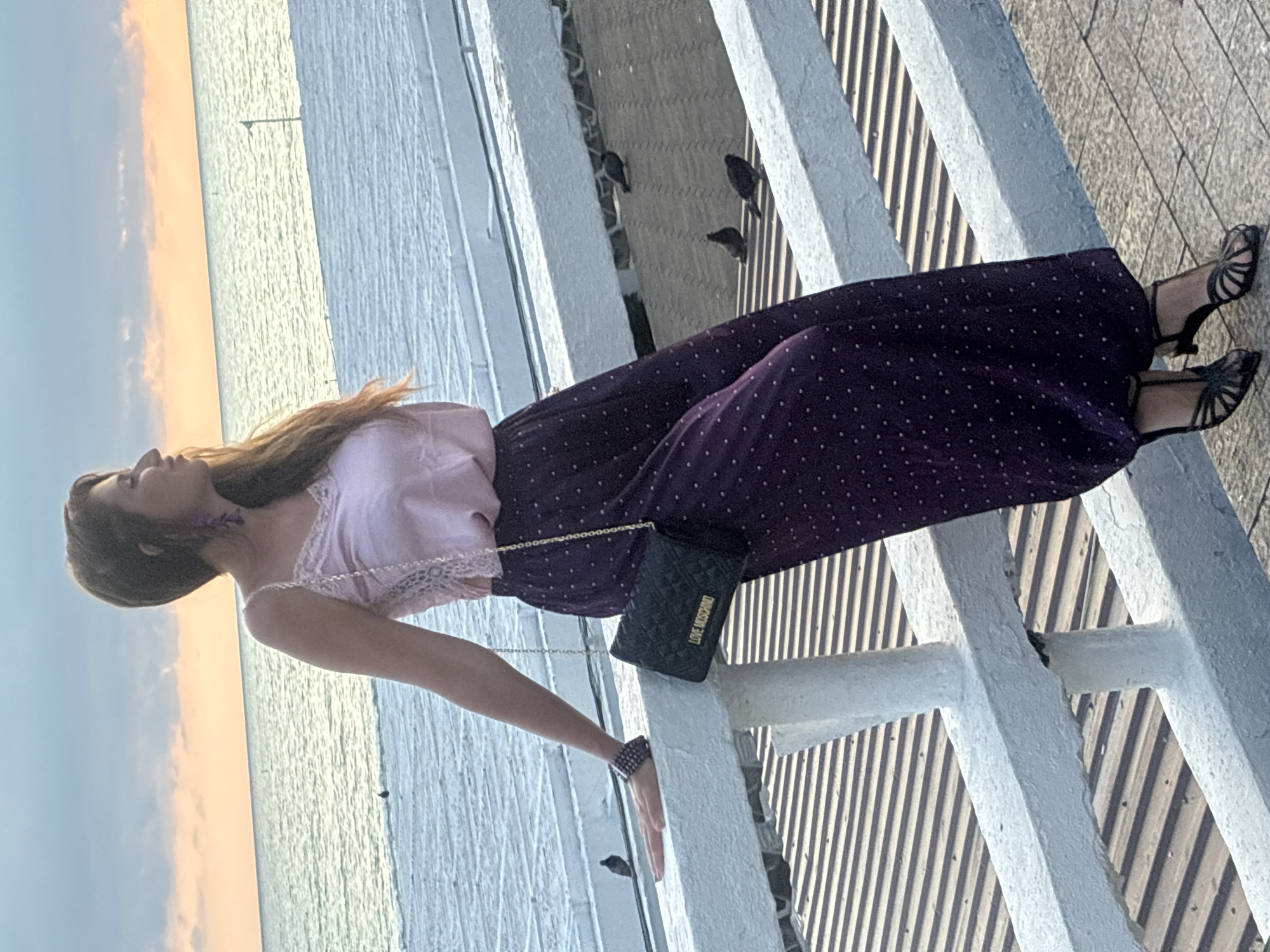 A woman is posing on a pier overlooking the sea at sunset. She is wearing a light pink camisole with lace trim, a dark purple polka-dot maxi skirt, and black open-toe heels. A black quilted crossbody bag with the "LOVE MOSCHINO" logo is slung across her body. She has long, auburn hair and is wearing dangling purple earrings. The pier is white and has a railing. Several pigeons are visible on the pier. The sky is a gradient of blue and orange, with the sun low on the horizon. The water is calm and reflects the colors of the sky. The location is the city of Sochi, Russia.
