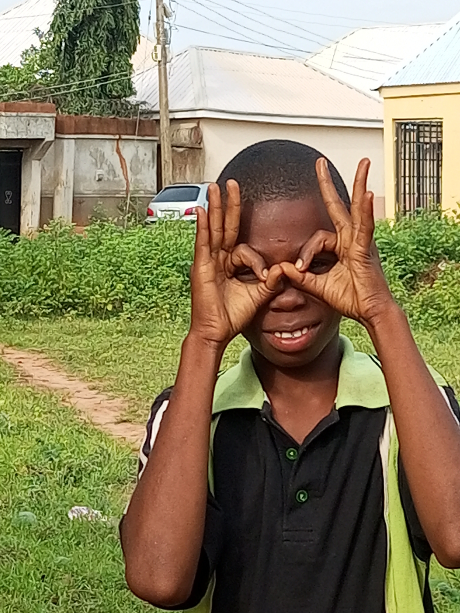 A young boy, likely of Nigerian descent, stands in a partially overgrown outdoor area that appears to be within a residential compound in Tempure, Nigeria. He is the central subject of the image, positioned in the foreground, and is looking directly at the camera with a playful expression. His hands are raised to his face, forming a circle around his eyes with his index fingers and thumbs, as if looking through binoculars or spectacles. He is wearing a black polo shirt with a green collar and white and green trim on the sleeves. The background features a light-colored building with a light blue roof, a smaller yellow building with barred windows, and a silver car parked in front of a brick wall. Dense green foliage, typical of the Nigerian climate, is visible throughout the scene, obscuring parts of the background and encroaching on a dirt path in the foreground. A utility pole with wires is also present to the left of the image. The lighting suggests it is daytime, with soft shadows indicating an overcast sky or late afternoon sun.