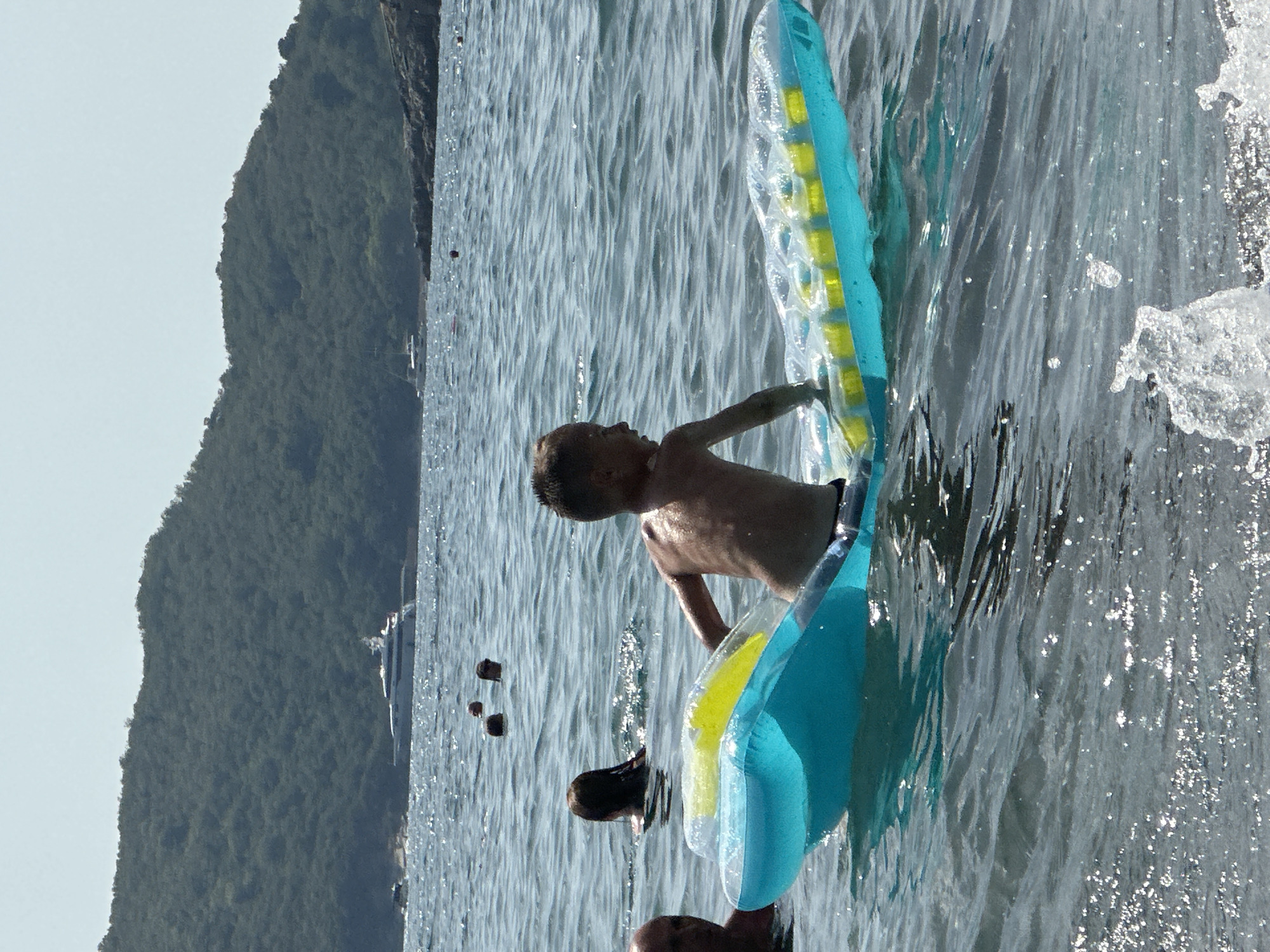 The image depicts a sunny day at the sea, likely near Boreti, Montenegro, as evidenced by the lush green hills in the background. In the foreground, a young boy is being propelled through the water on a blue and yellow inflatable raft. He is looking to his left, his skin glistening with water. To the left of the raft, another person's head and shoulders are visible, partially submerged in the water. Further in the background, several other people are swimming or floating in the sea. In the distance, a large white yacht is anchored near the coast, and smaller boats can be seen closer to the shore. The water is a deep blue, with gentle ripples and sunlight glinting off its surface. The overall scene conveys a sense of leisure and summer enjoyment.