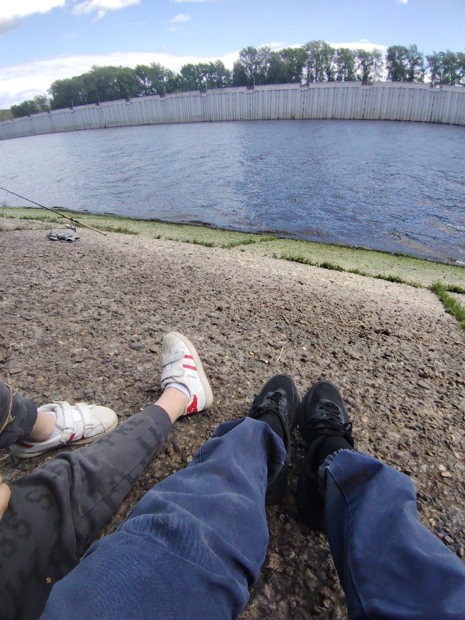 Two people are sitting on a concrete embankment by a river or body of water. A fishing rod and tackle are visible nearby. The embankment is lined with green vegetation where it meets the water. In the background, a long concrete retaining wall is visible behind trees. The first person wears grey sweatpants and sneakers with white and red stripes. The second person wears blue jeans and black sneakers. The location is likely in the city of Chaikovsky, Russia.