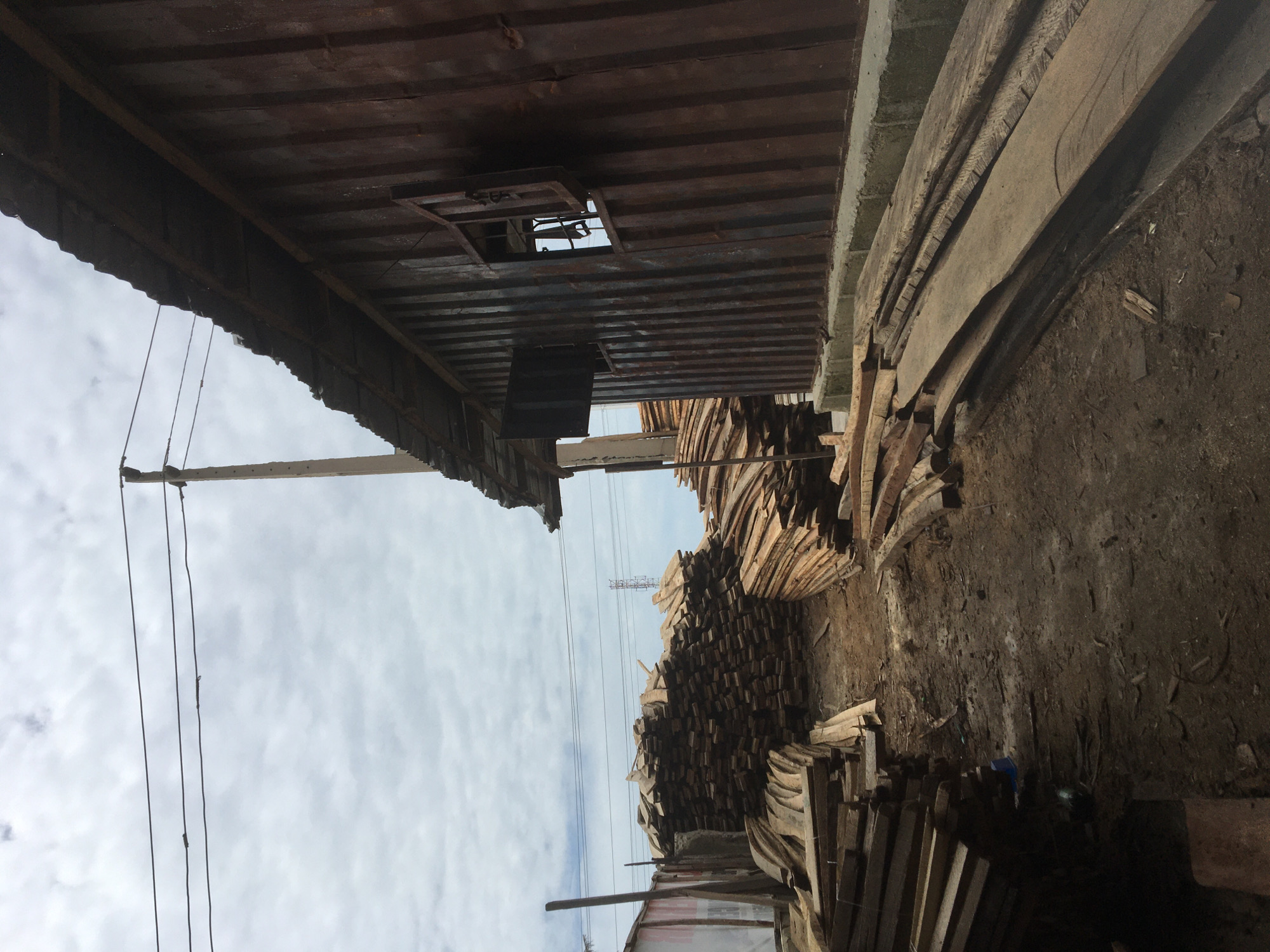 This image, taken in Ado, Nigeria, depicts an outdoor lumber or wood storage facility under an overcast sky. The foreground is dominated by the dark, corrugated metal underside of an overhead structure, possibly a shed roof. An opening, framed by metal, is visible within this structure.
Below, the midground is filled with numerous stacks of rough-hewn wooden planks and boards of varying lengths and widths. Some stacks are neatly organized into rectangular piles, while others are arranged less formally, including some in curved configurations. The ground beneath the wood piles is uneven, dark dirt, with scattered debris.
To the left, a utility pole or wooden beam stands vertically, supporting several dark wires that stretch across the upper portion of the frame. The sky, visible above the wooden stacks and wires, is covered by a dense layer of white and gray clouds, indicating daytime. In the distant background, silhouetted against the sky, are multiple power lines and a large metal power transmission tower.
No people are present, and no active events are occurring; the scene captures a static storage area. There is no visible text. The overall atmosphere suggests an industrial or semi-industrial environment focused on wood processing or storage.