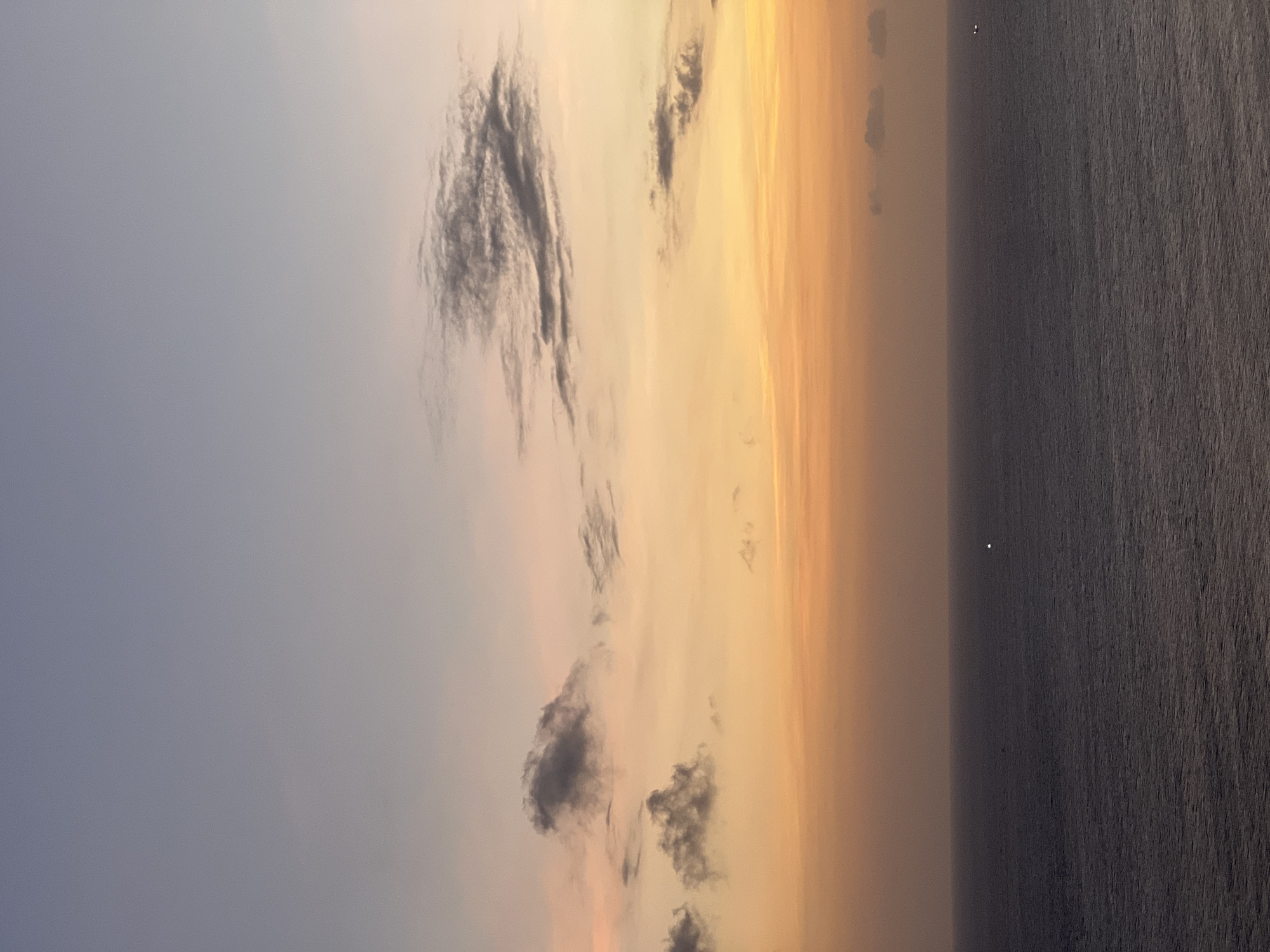 The image shows a sunset view from Kuta Selatan, Indonesia. The sky transitions from a pale blue-gray to a vibrant orange-yellow near the horizon. Dark gray clouds are scattered throughout the sky. The ocean is dark and appears calm, stretching horizontally to the right edge of the image. A single, bright point of light, possibly a star or distant light source, is visible over the ocean.