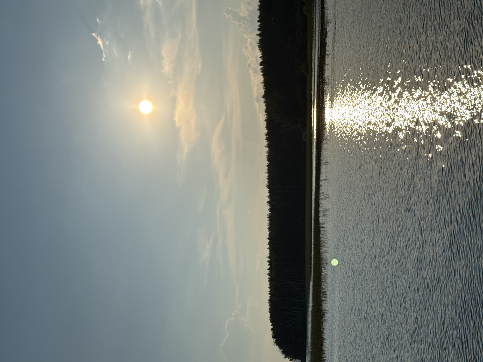 The image depicts a serene landscape scene, likely a waterfront area, with a focus on natural elements. The sky is a pale blue, transitioning to a lighter hue near the horizon, with scattered, wispy clouds. A bright, circular sun is positioned in the upper right quadrant of the sky, emitting a soft glow and creating a reflection on the water. Below the sky, a dense line of dark green trees forms a treeline that stretches across the middle ground, suggesting a forest or a heavily wooded area. In front of the treeline, a body of water, likely a river or lake, dominates the lower half of the image. The water is rippled, and a prominent, bright reflection of the sun sparkles across its surface, creating a path of shimmering light. There is also a faint, green circular object visible on the water's surface, to the left of the sun's reflection. The foreground shows the textured surface of the water. The overall impression is one of natural beauty and tranquility, with the sun and its reflection being a key visual element. This scene is consistent with locations along the Kama River in Chaykovsky, Russia, known for its picturesque riverbanks and surrounding forests.