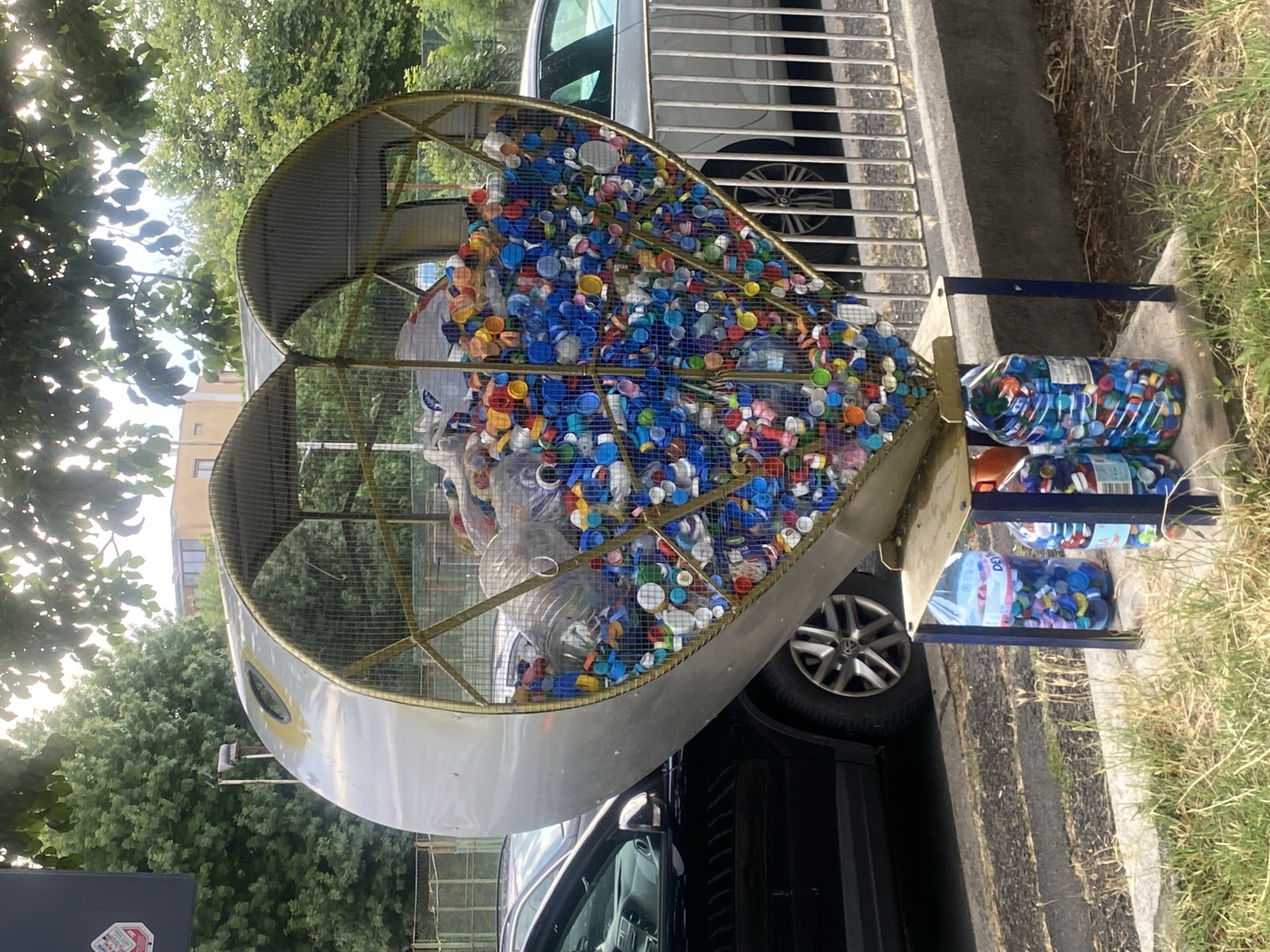 The image shows a large, heart-shaped recycling bin overflowing with plastic bottle caps of various colors. The bin is made of metal and has a mesh interior. It is supported by a blue metal stand. Below the bin, two large plastic bottles are filled with bottle caps, serving as further collection points. The recycling bin and bottles are situated outdoors, with a black car parked to the left and a silver car visible behind a metal fence to the right. Trees and some buildings are in the background. The scene appears to be in an urban environment. This specific recycling bin is located in Burgas, Bulgaria, near the entrance to the Sea Garden.