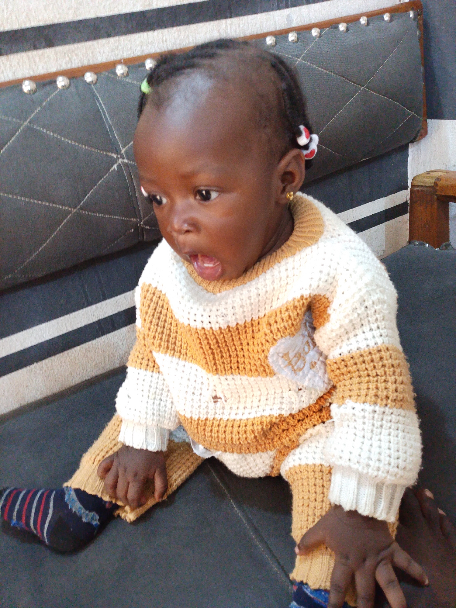 The image shows a close-up of a young child, likely a toddler, sitting on a dark-colored surface. The child has dark skin and is wearing a knitted sweater with horizontal stripes of white and orange-brown. The sweater has a high neckline. On the child's head, there are braided pigtails adorned with small, colorful beads. The child is also wearing a dark sock with vertical stripes of blue, red, and white. The background consists of a striped wall and a piece of furniture, possibly a sofa or chair, with decorative studs. The child's expression appears surprised or excited, with their mouth open. The lighting suggests it is daytime. There is no visible text in the image. The context provided indicates the location is Jos, Nigeria.