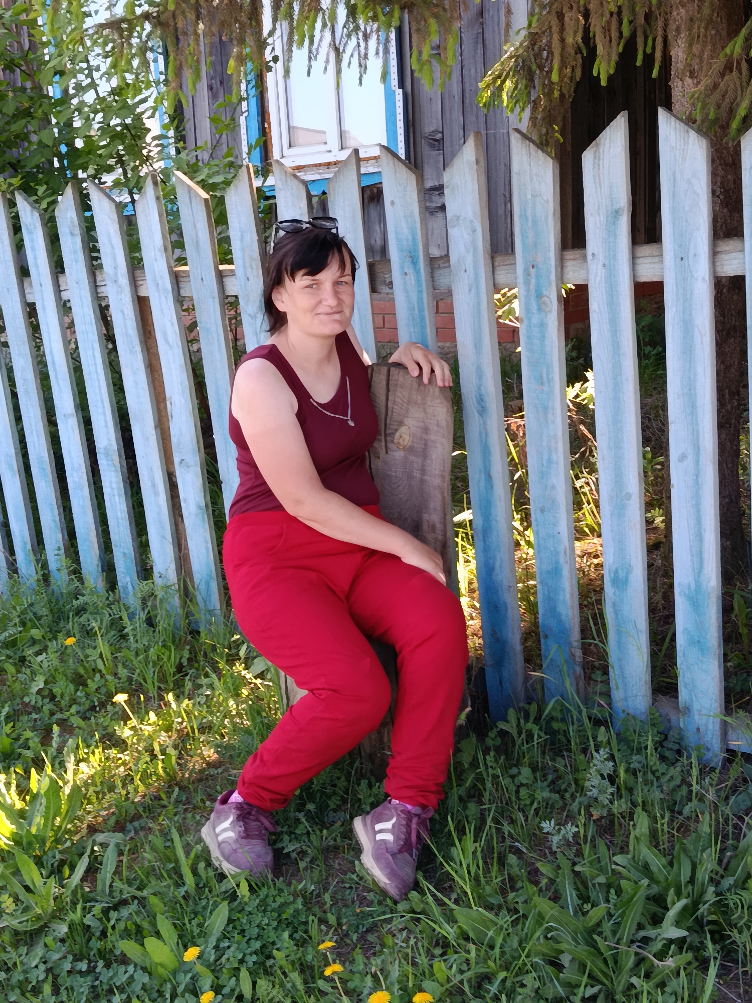 A woman is seated beside a faded blue wooden fence in Фоки, Russia. She wears a maroon sleeveless top, red pants, and purple sneakers. Sunglasses are placed on top of her head. The ground is covered in grass and dandelions. A wooden structure or post is next to her, and part of a building is visible behind the fence.