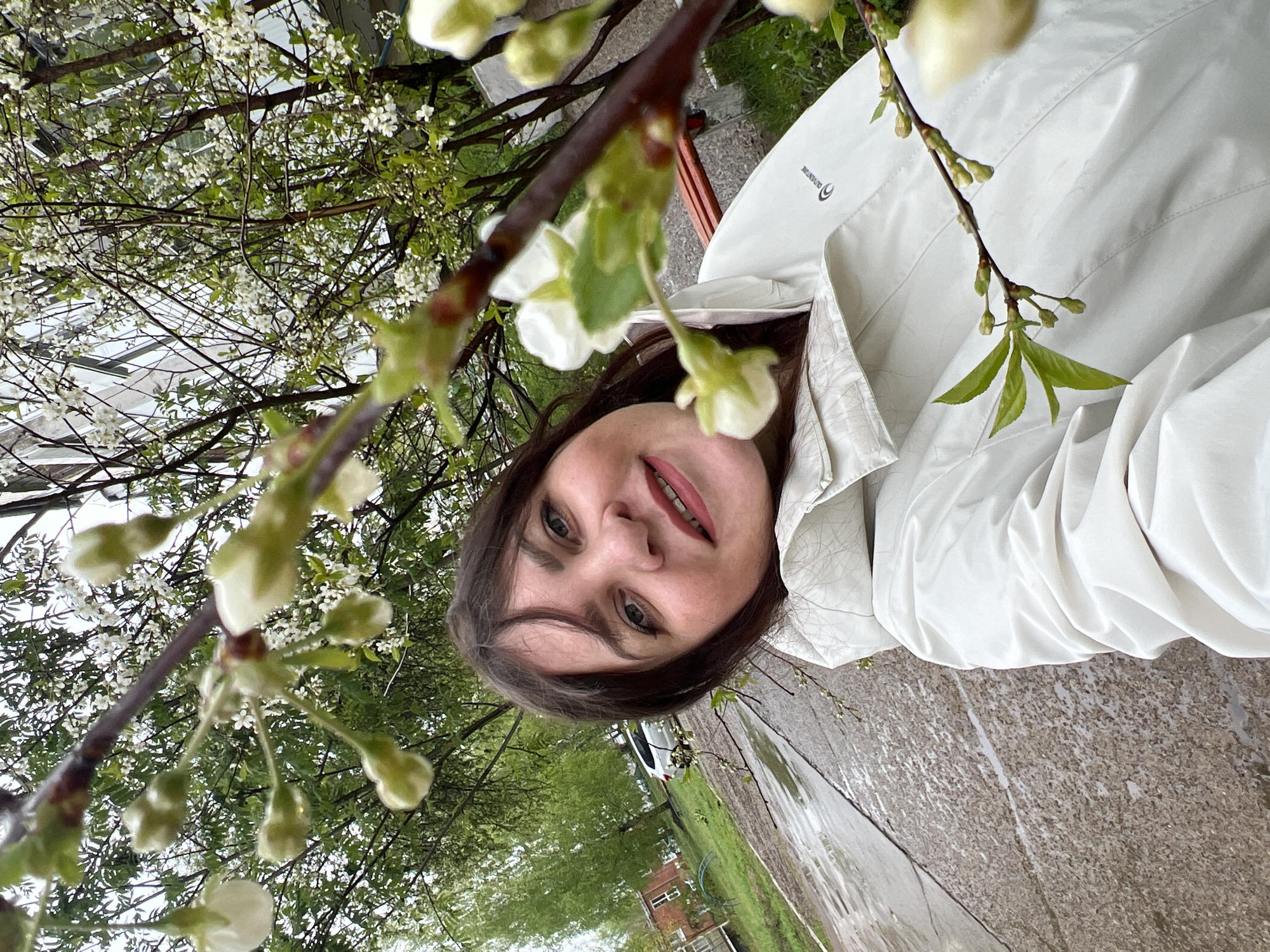 A woman with brown hair and fair skin stands outside, possibly on a sidewalk in Noviy, Russia. She is wearing a light-colored jacket. The background shows a rainy street, trees, grass, and buildings. A branch with white buds is close to the camera, partially obscuring the view. The image is rotated counterclockwise.