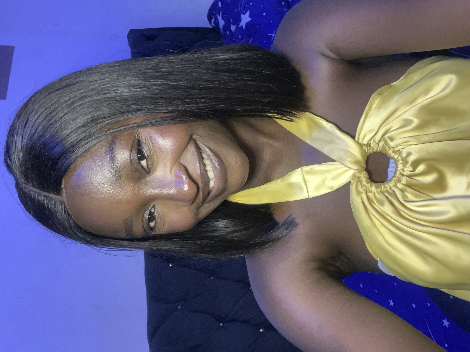 A dark-skinned woman with short black hair is lying on a dark blue, quilted surface, possibly a bed or a sofa. She is wearing a yellow, silky halter top. The backdrop includes a blue surface, likely a wall, and a blue blanket with white stars on it. The overall lighting appears to be artificial. No information about Jos, Nigeria, is identifiable in the scene.