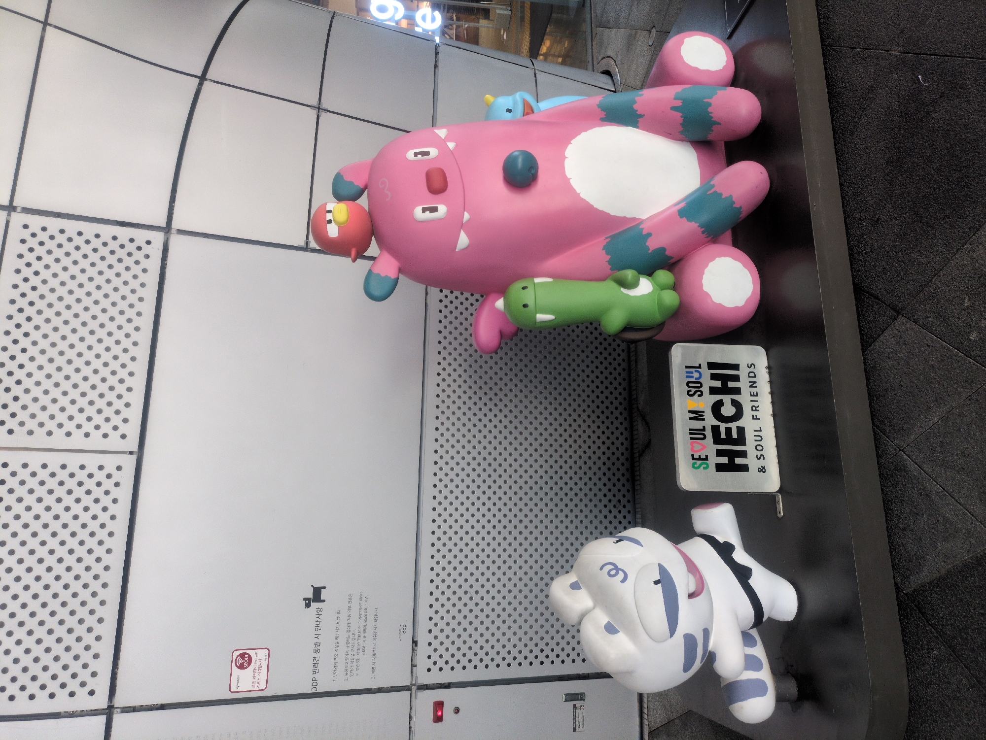 The image shows colorful cartoon character sculptures. A pink, cartoon monster is on its back, with a green cartoon animal lying on its stomach. Next to it is a sign that reads "SEOUL M SOUL HECHI & SOUL FRIENDS". Below the monster is a white and blue cartoon character. The background appears to be a gray tile floor and a light gray wall. A sign on the wall is written in Korean and includes dog guidelines. The location is likely a public space in Seoul, South Korea, as indicated by the signs.