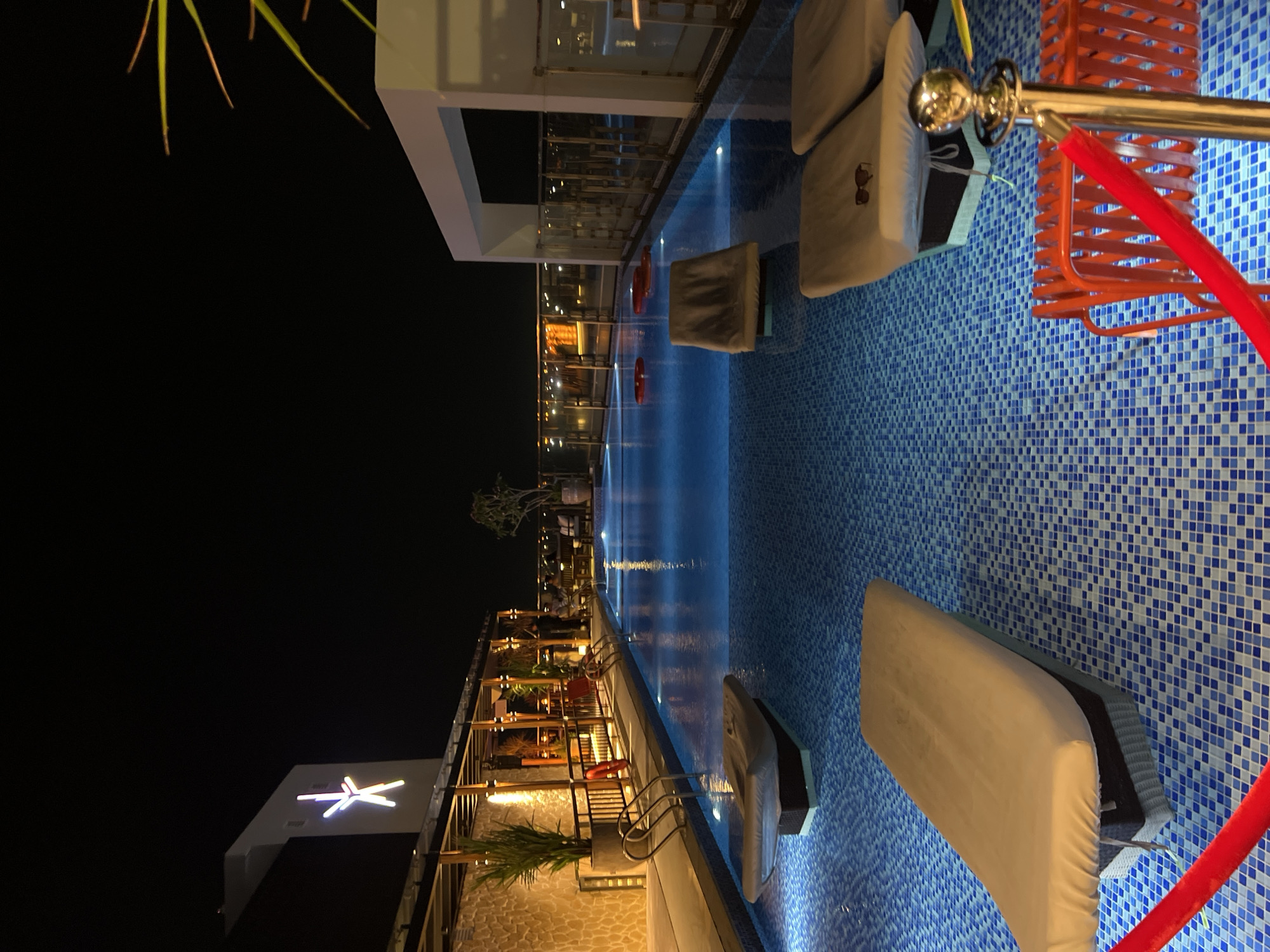The image shows an outdoor swimming pool area at night. The pool is lined with blue and white mosaic tiles and has several lounge chairs around it, some of which are covered with white towels. The pool reflects the lights from nearby buildings, creating a shimmering effect. An orange chair and gold pole with a red rope are also visible in the foreground. The scene is located in Nha Trang Ward, Vietnam.